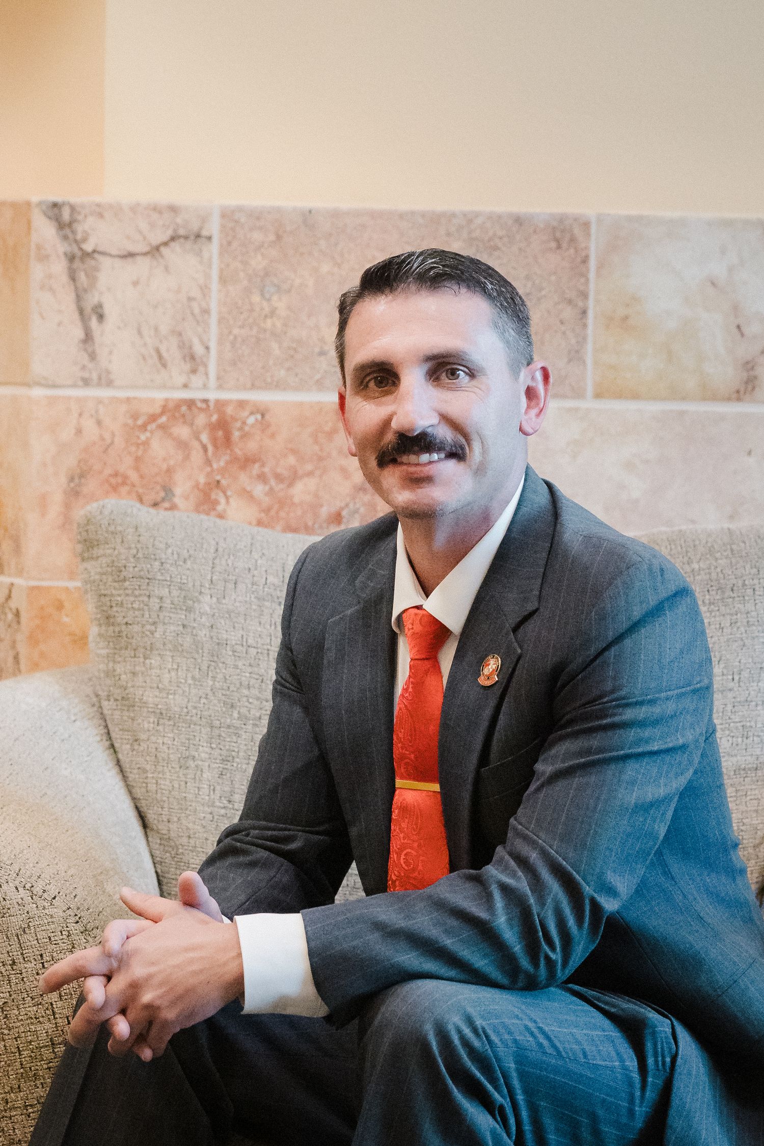 Man in gray suit headshot — Professional man in a gray suit and orange tie sitting in a modern lobby setting.