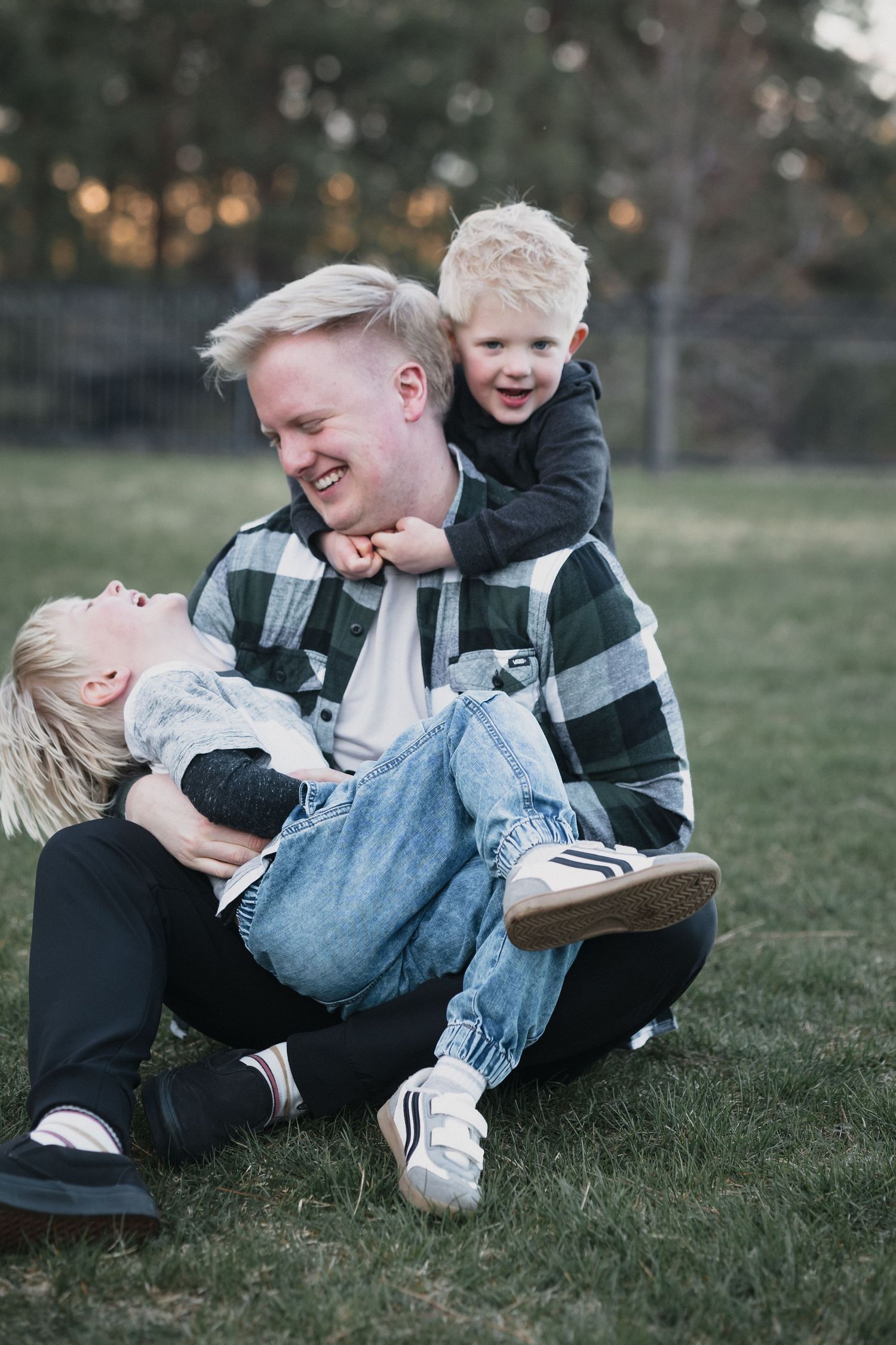 Laughing father playing with two energetic young blonde boys while sitting on green grass.