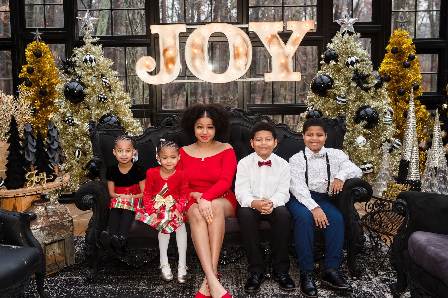Family of five posing joyfully in front of a festive JOY sign with Christmas trees during holiday photo session.
