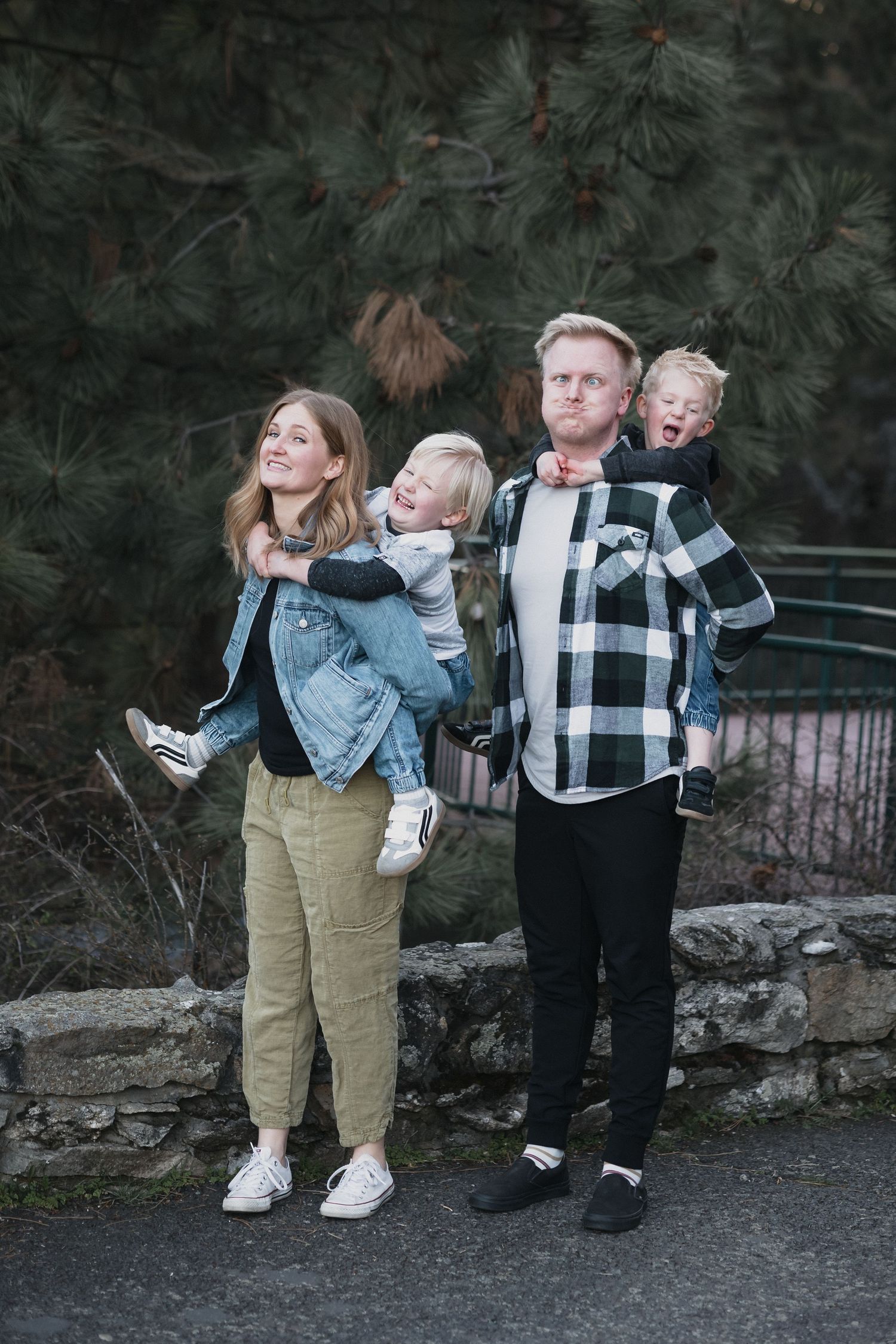 Family of four stone wall — Family of four posing outdoors as parents hold their two young blonde children near a stone wall and pine trees.