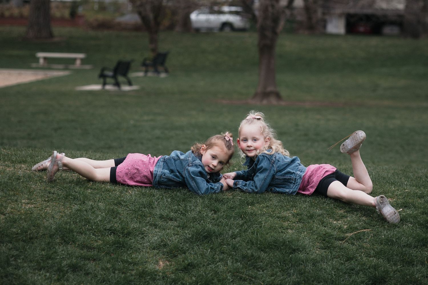 Two girls denim jackets grass — Two young girls in denim jackets lying on green grass in a park, smiling together.
