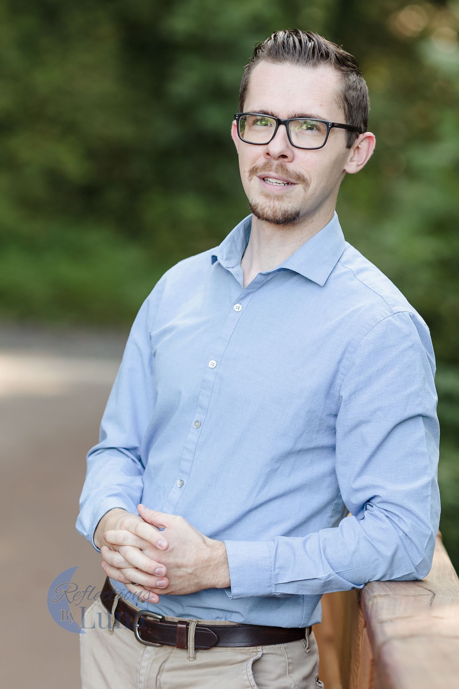 Hands clasped portrait — Confident outdoor portrait of man in blue shirt and glasses standing with hands clasped and smiling.