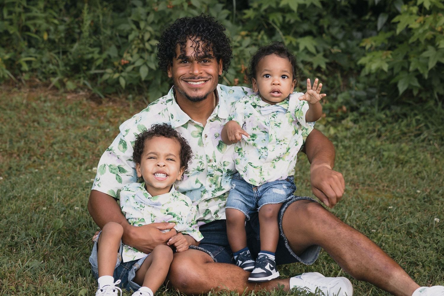 A man and two young children wearing matching floral shirts sit together smiling on green grass outdoors.