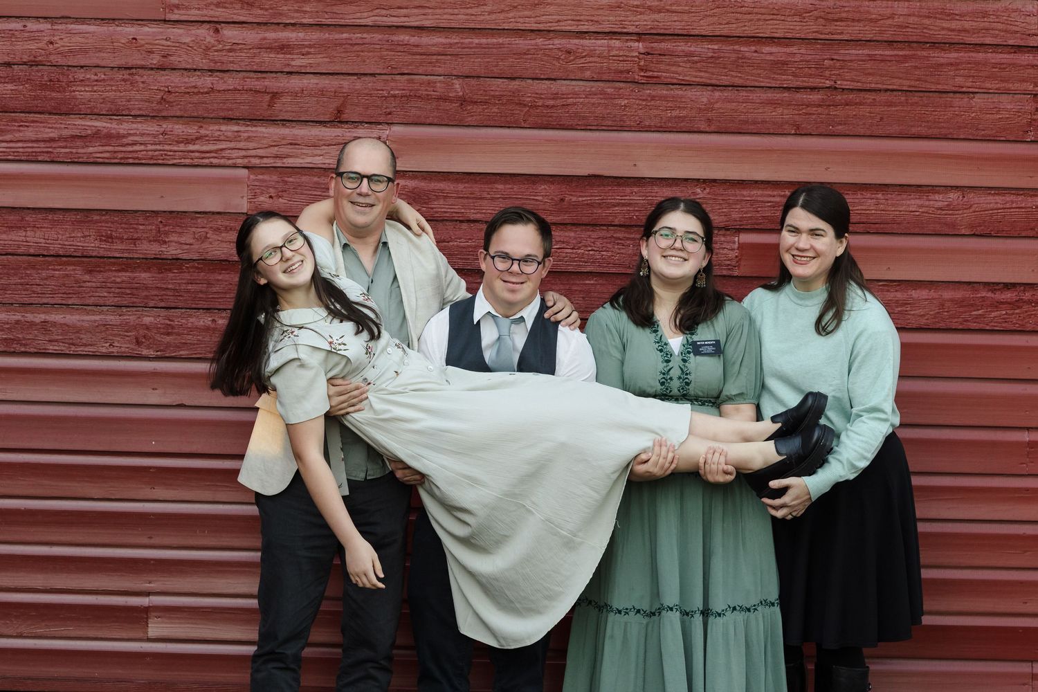 A joyful family group photo with five people posing and laughing in front of a red barn wall.