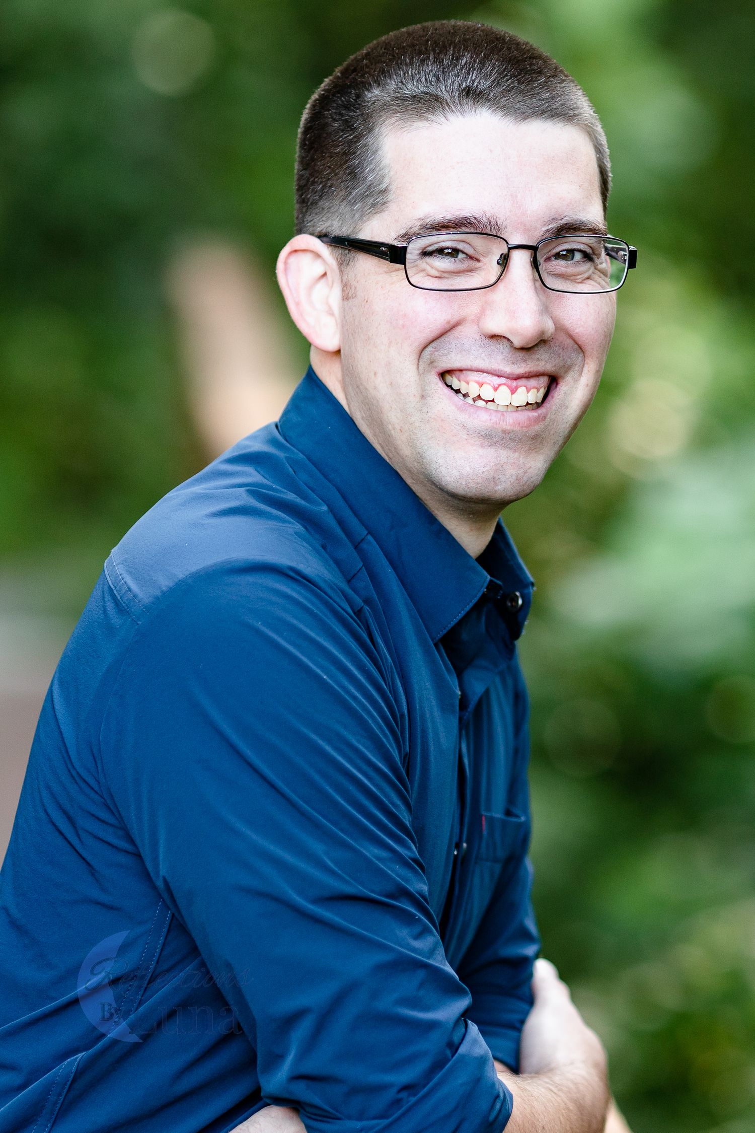 Leaning on railing headshot — Relaxed professional headshot of man in blue shirt and glasses leaning on wooden railing outdoors.