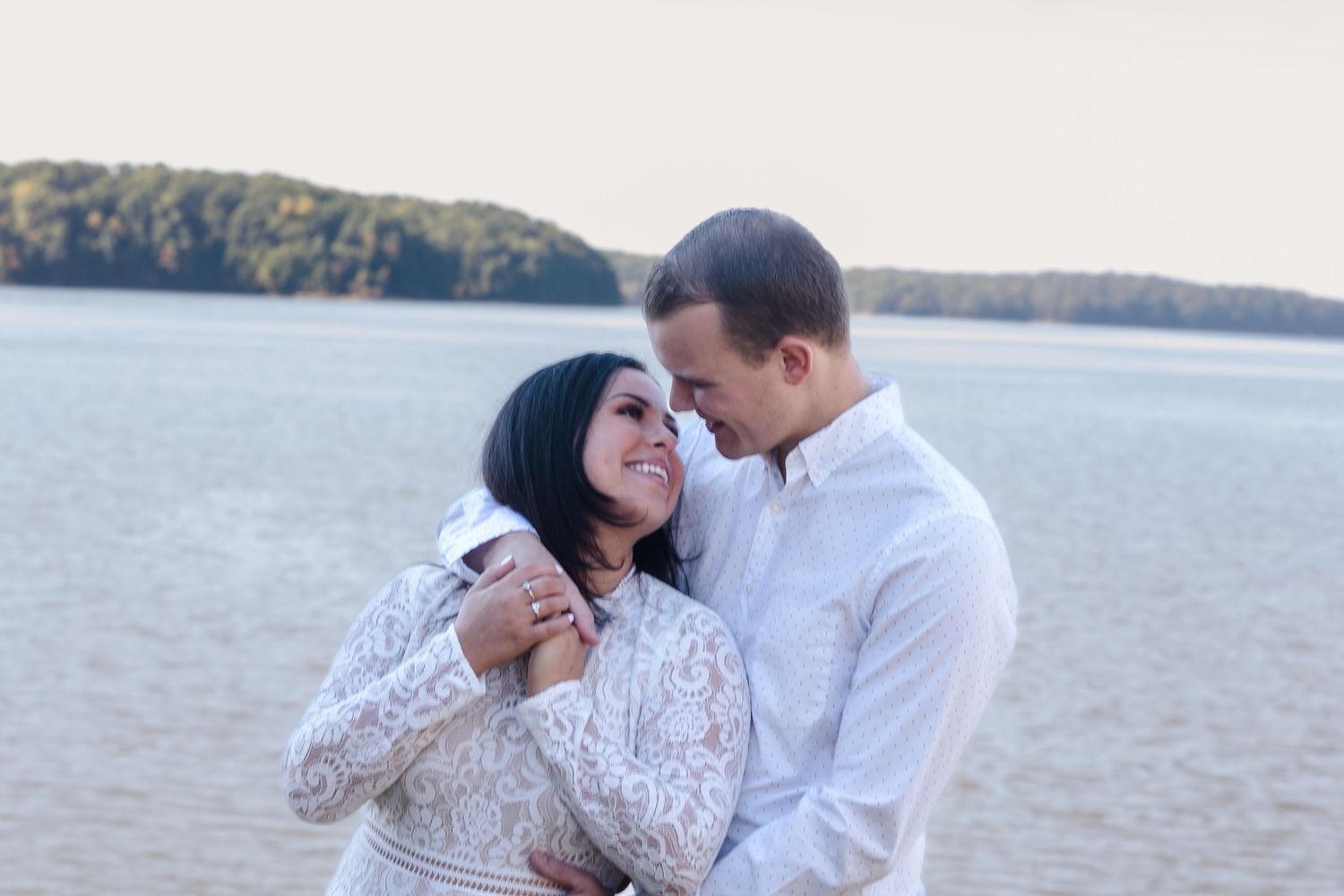 Happy couple embracing and smiling on sandy beach in matching white outfits during engagement photo.