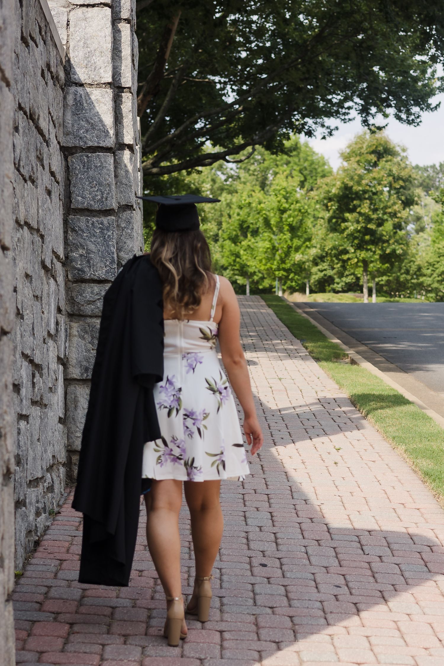Graduate in floral dress carrying cap and gown walks along brick path beside stone wall.