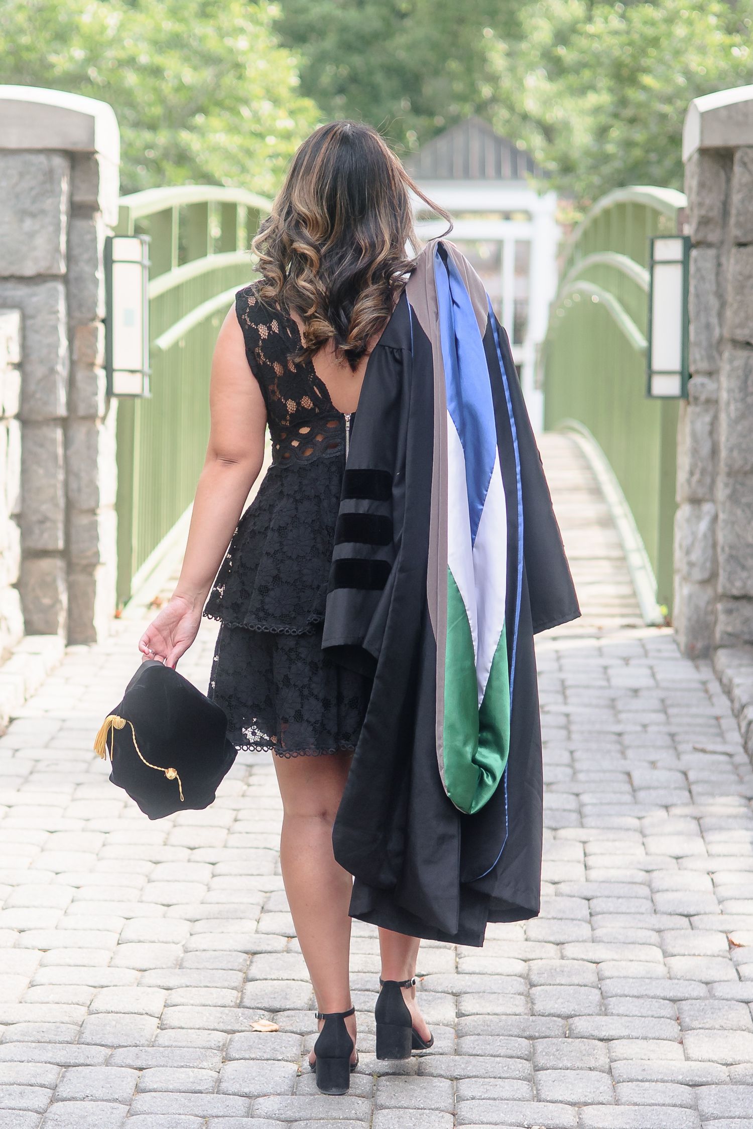 Graduate in black lace dress carries cap and gown while walking toward green bridge entrance.