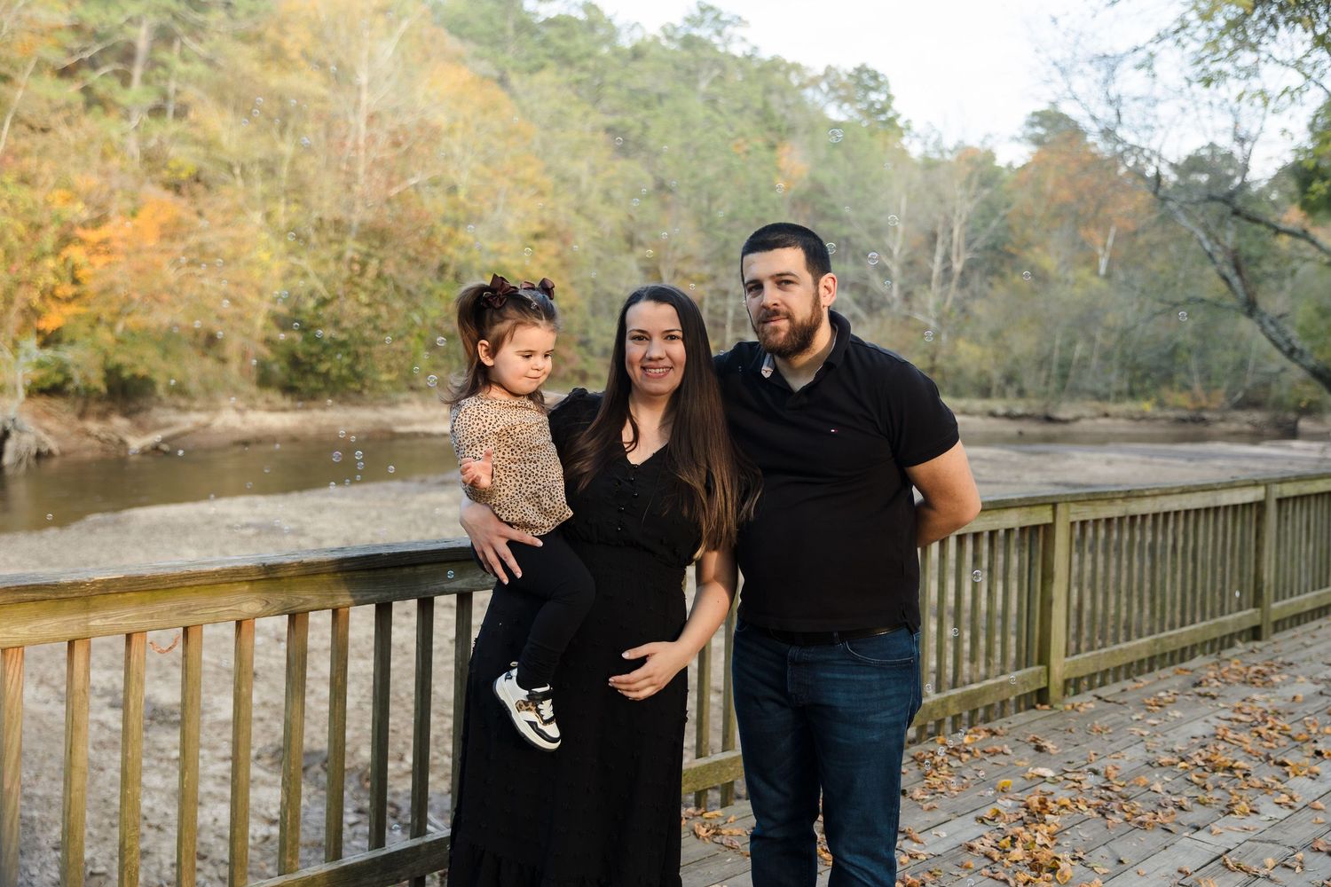 Pregnancy family on bridge — Fall family portrait of pregnant woman in black dress posing with partner and toddler on wooden bridge.