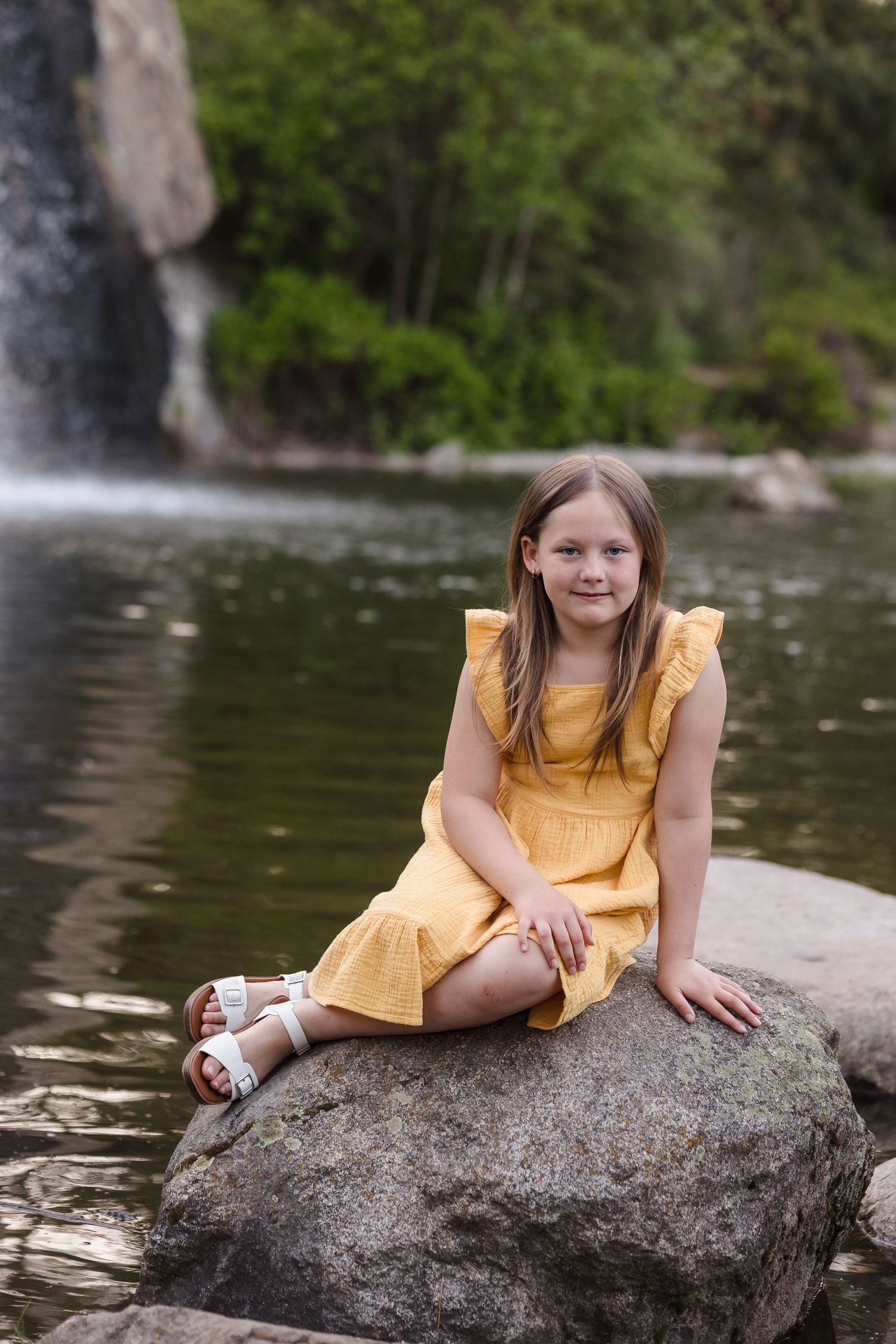Young girl in yellow dress sitting on a rock by a waterfall in a lush green park, smiling at the camera.
