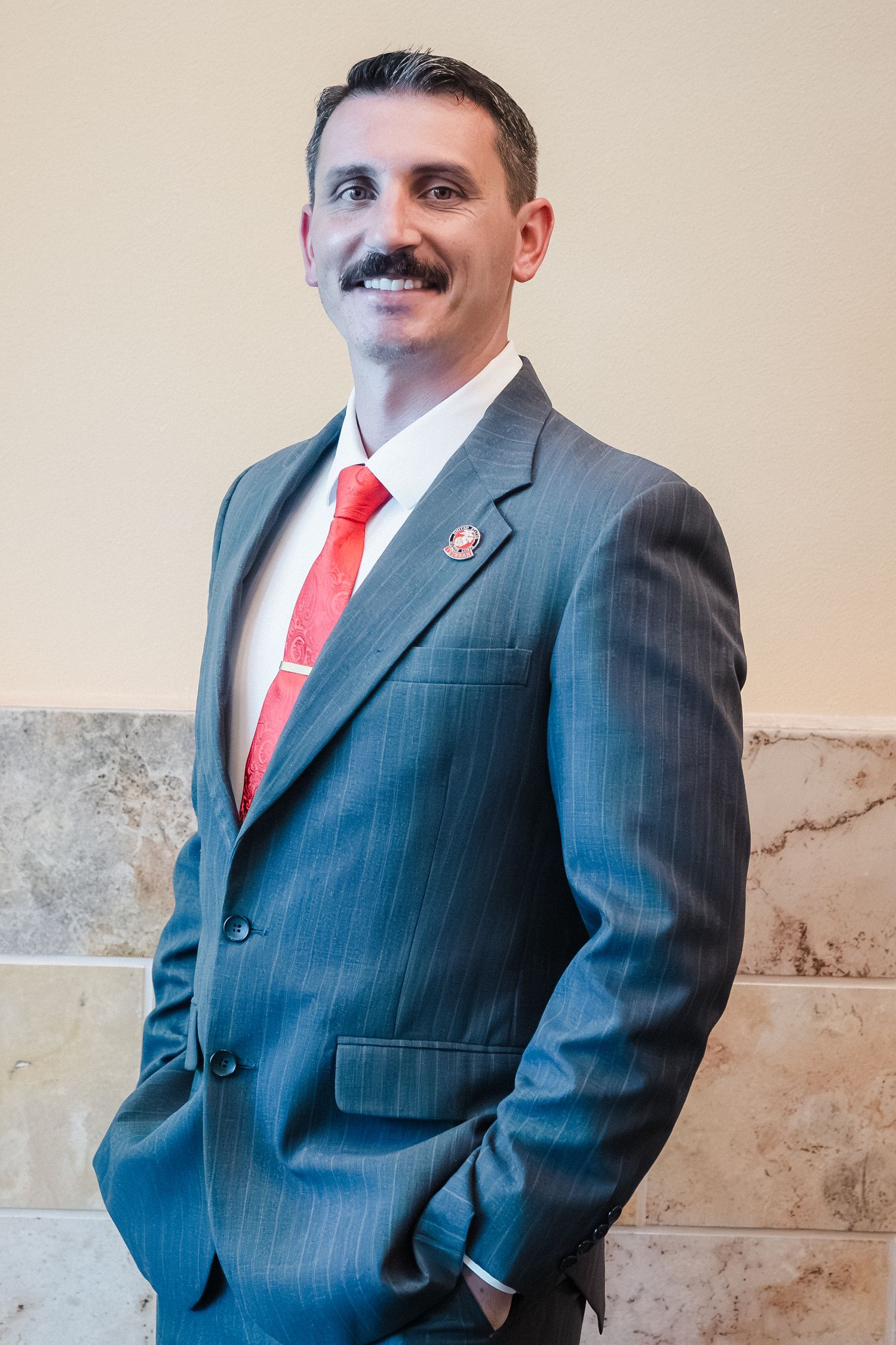 Professional man with mustache wearing gray pinstripe suit and red tie standing in marble hallway.
