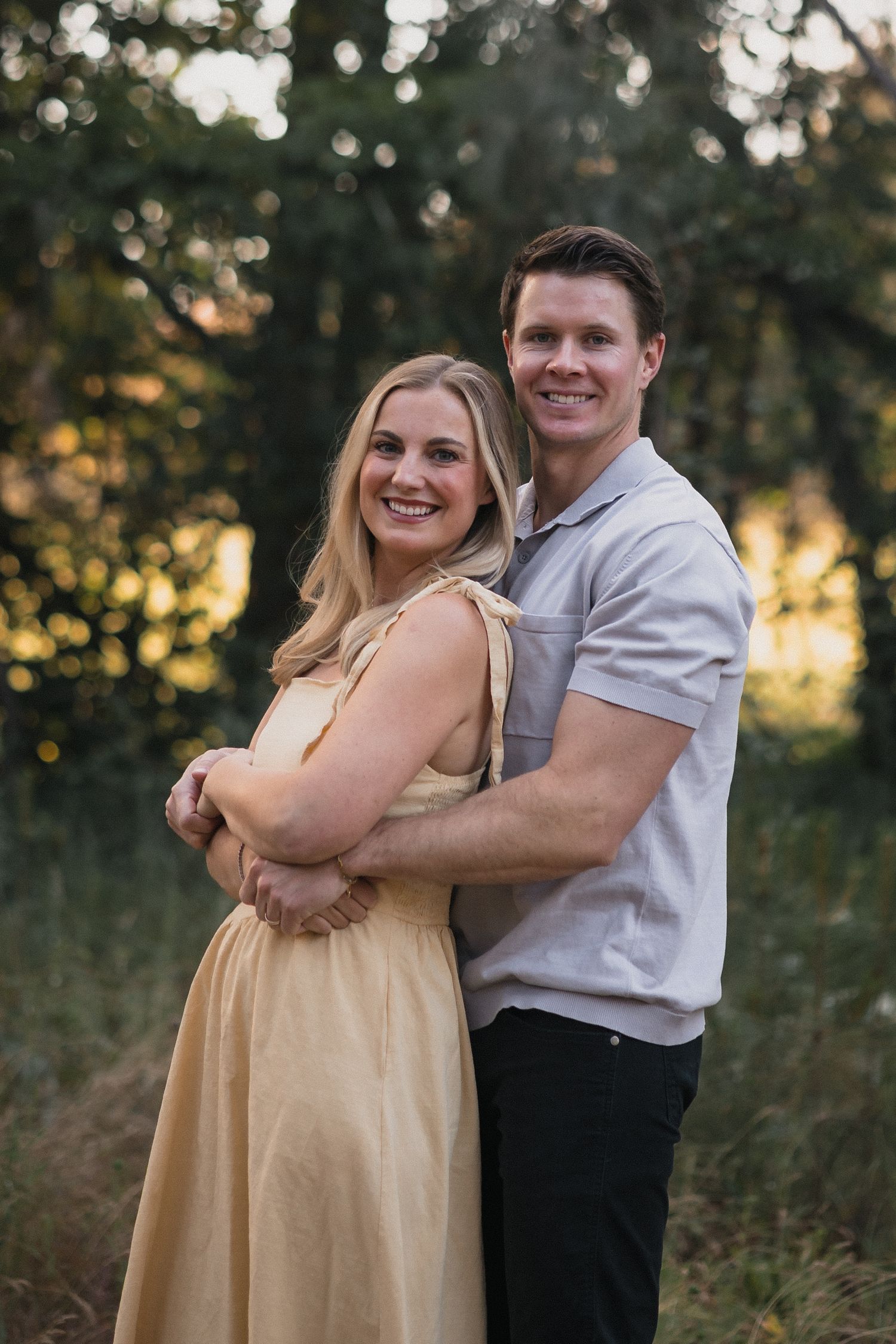 A smiling couple embraces outdoors at golden hour, woman in yellow dress, man in gray polo, with lush green trees behind them.