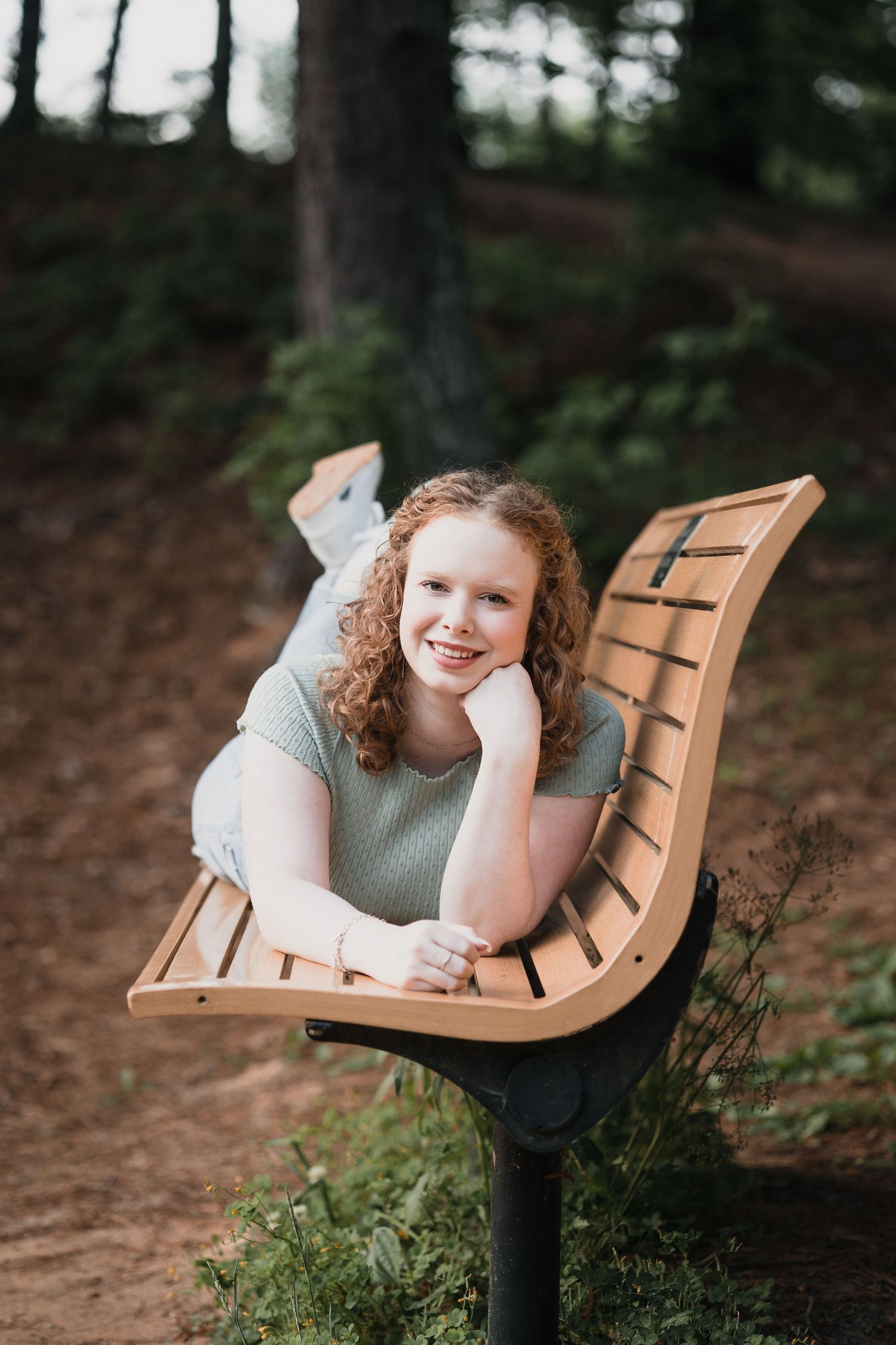 Red‑haired senior on bench — Curly red‑haired teen girl lying on a modern wooden bench in a pine forest, smiling at the camera.