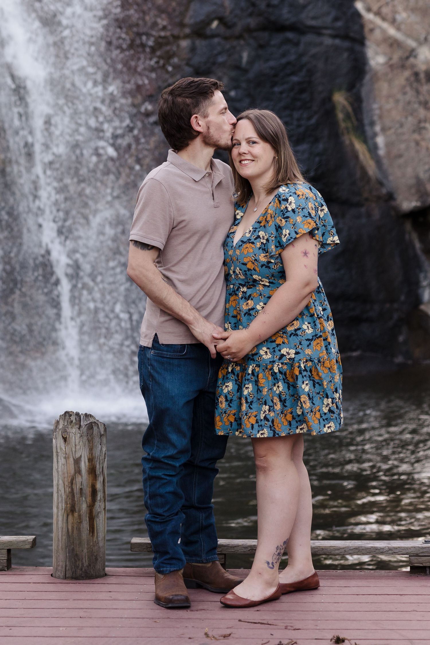 Couple by waterfall dock — Couple holding hands in front of a waterfall as he kisses her forehead on a wooden dock.