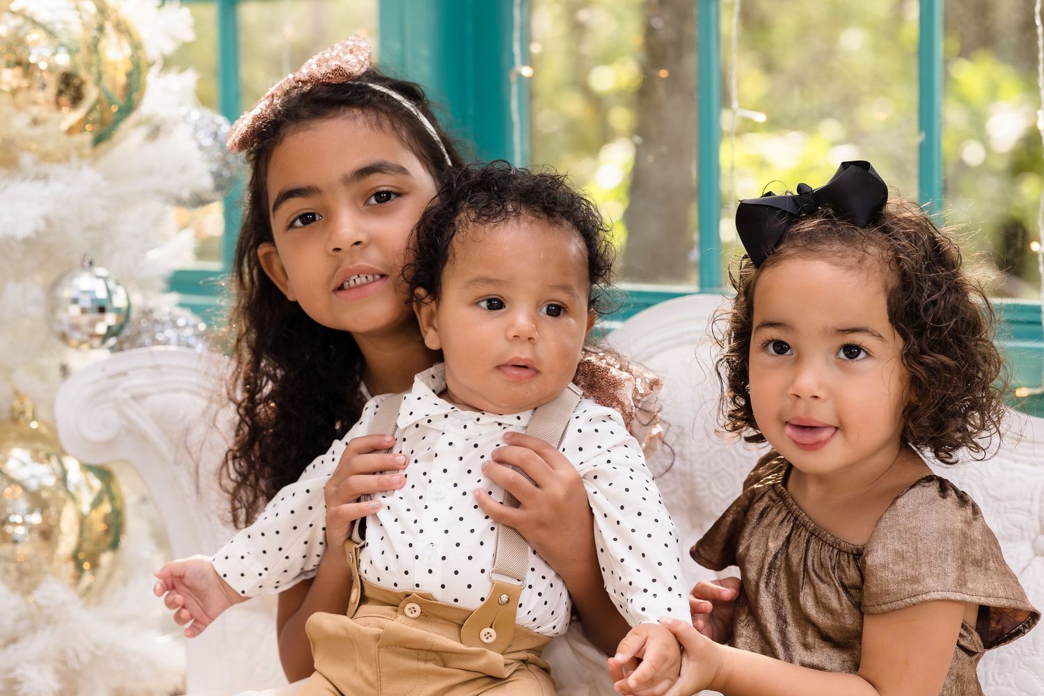 Three young children pose together near a white Christmas tree in a festive holiday portrait setting.