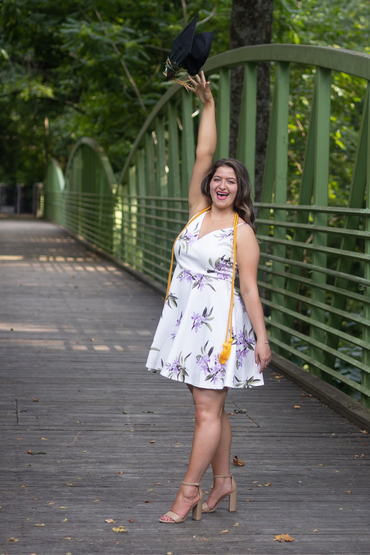 Joyful graduate in floral dress tosses cap in air on green bridge surrounded by trees.