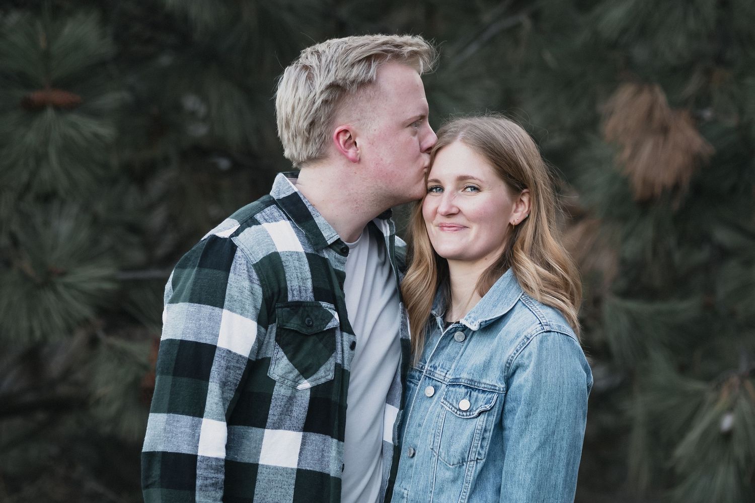 Couple kissing by pines — Young couple sharing a tender kiss near evergreen pine trees during an outdoor session.
