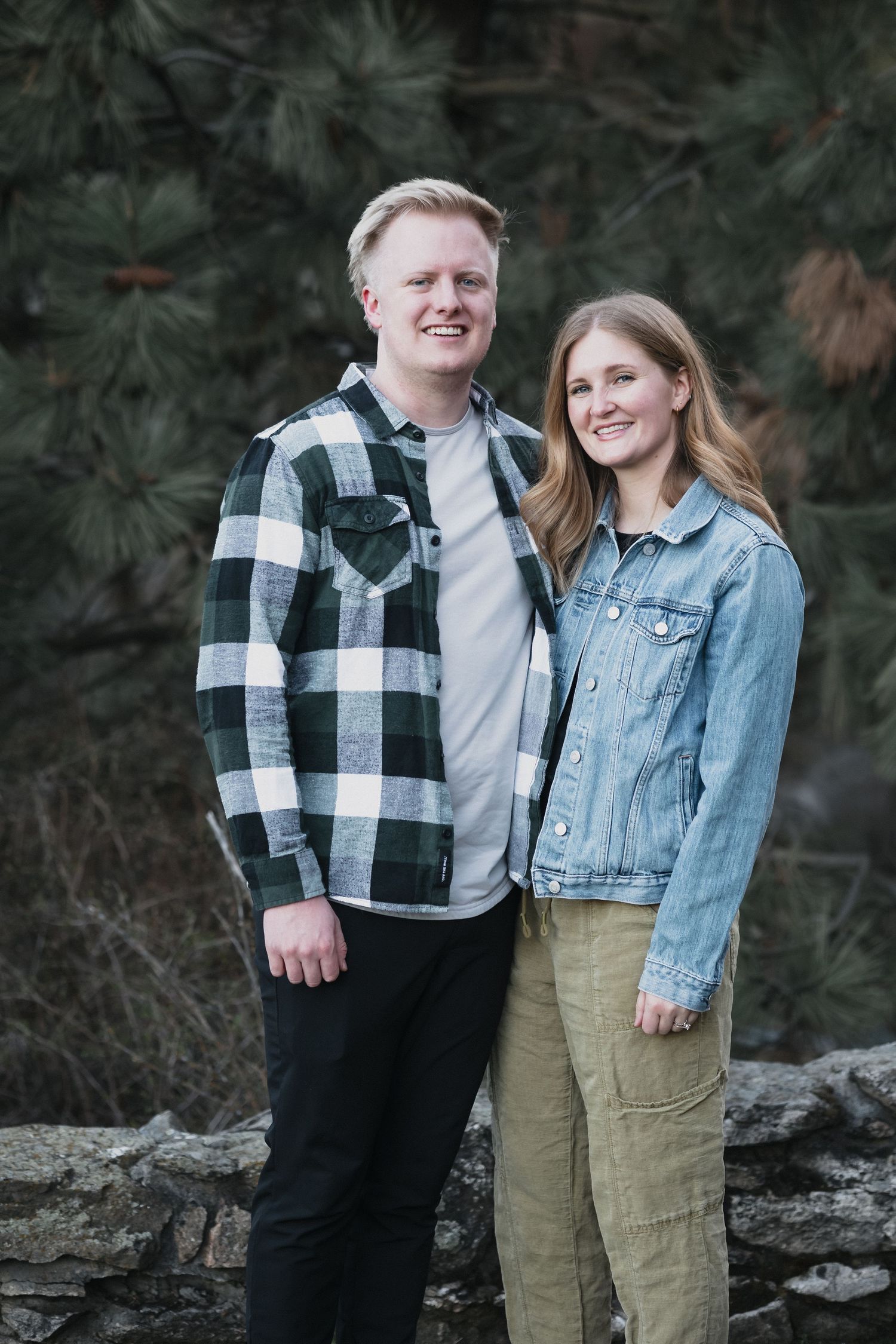 Couple pine trees stone wall — Smiling couple standing together outdoors in front of pine trees and a stone wall.