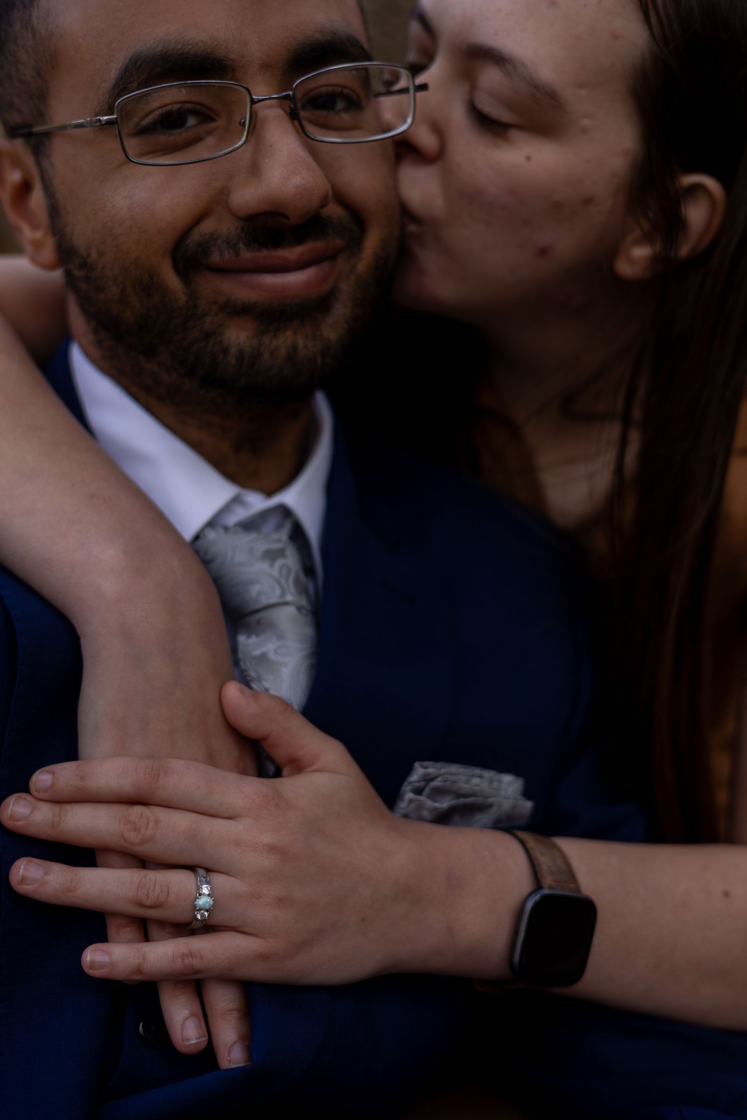 Engagement cheek kiss — Woman kissing her fiancé’s cheek while hugging him from behind, showing her engagement ring as he smiles.