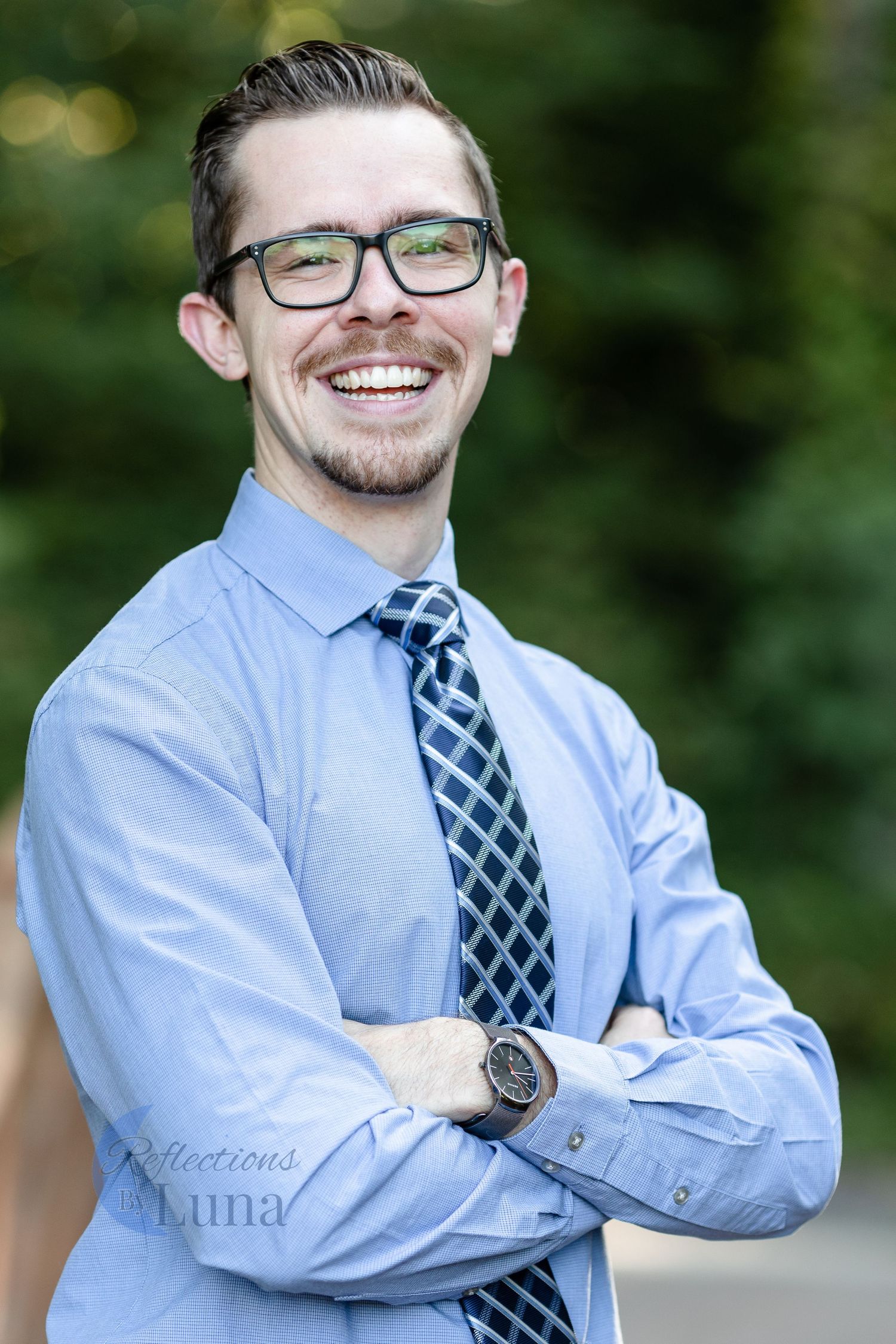 Outdoor headshot arms crossed — Professional outdoor headshot of smiling man in blue shirt and glasses with arms crossed in natural light.