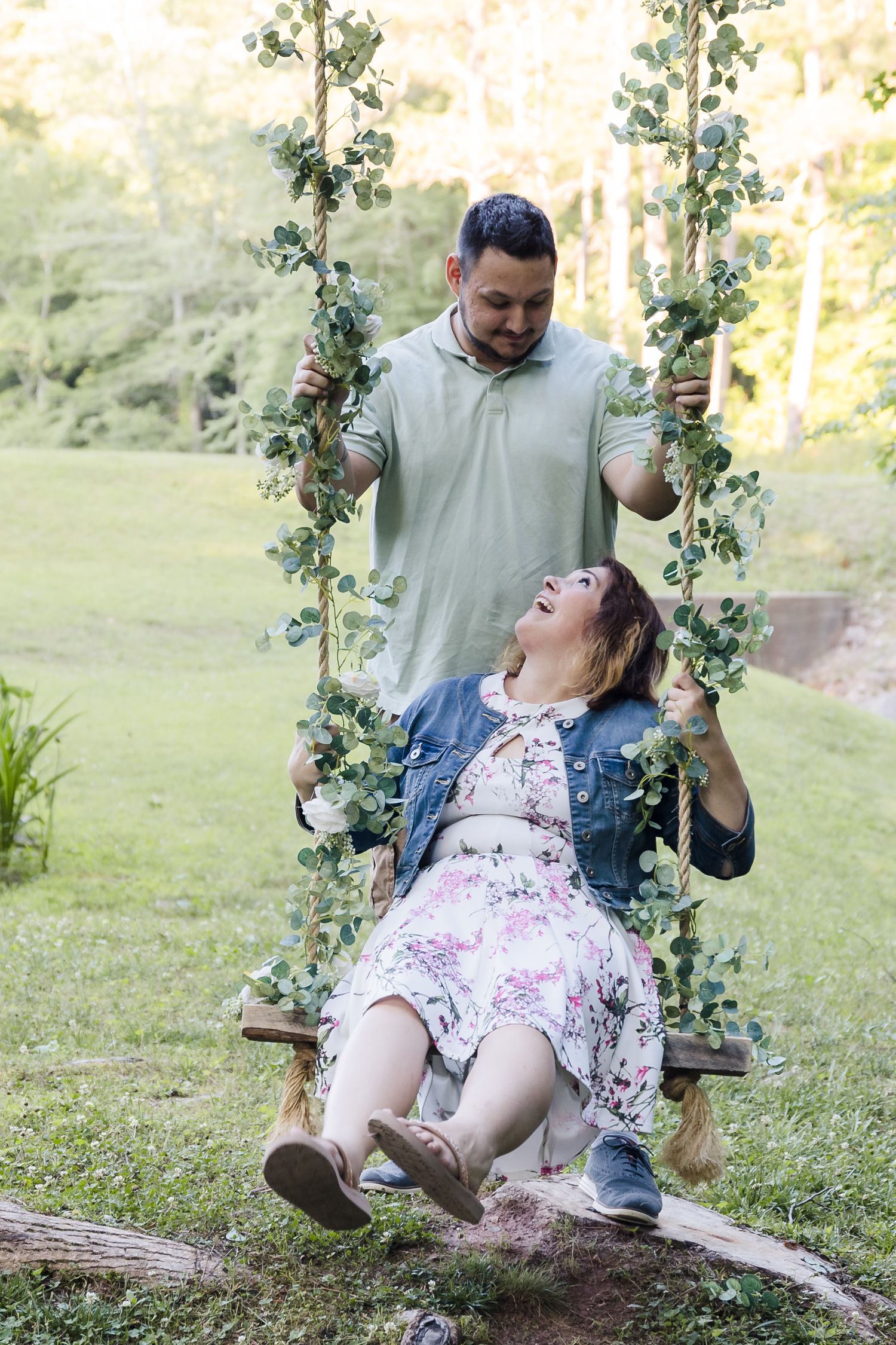 Garden swing couple — Man pushing a laughing woman on a eucalyptus‑decorated garden swing in a lush green park.