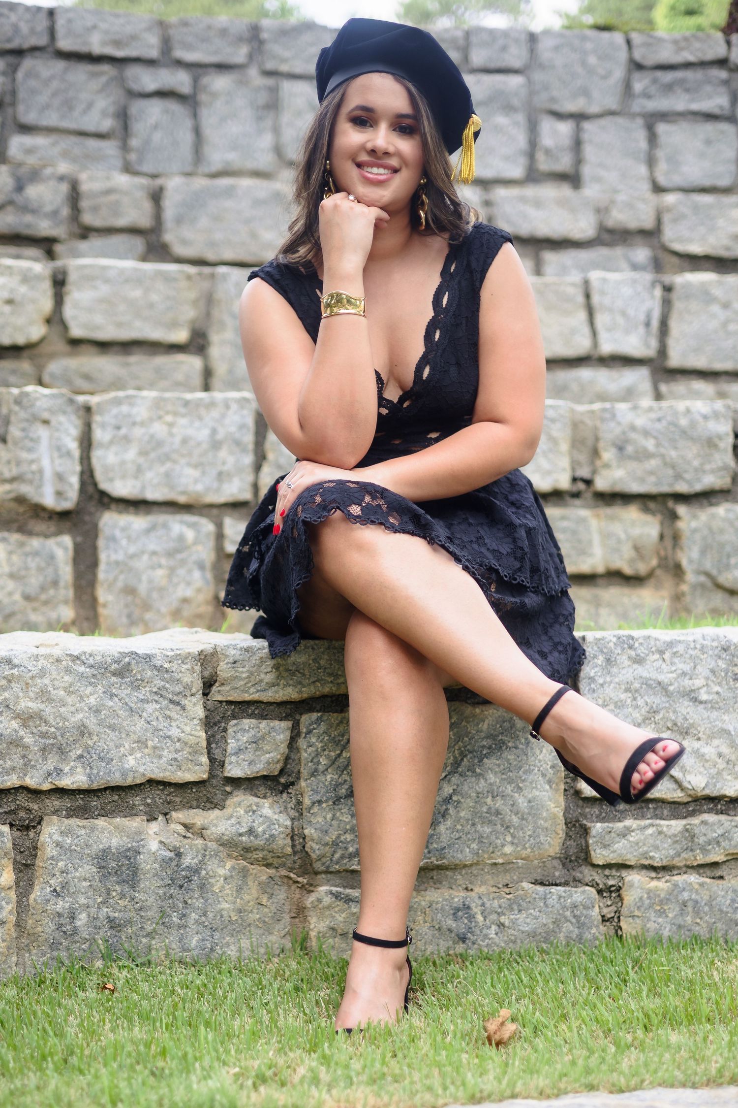 Smiling graduate in black lace dress and cap sits on stone wall with legs crossed.