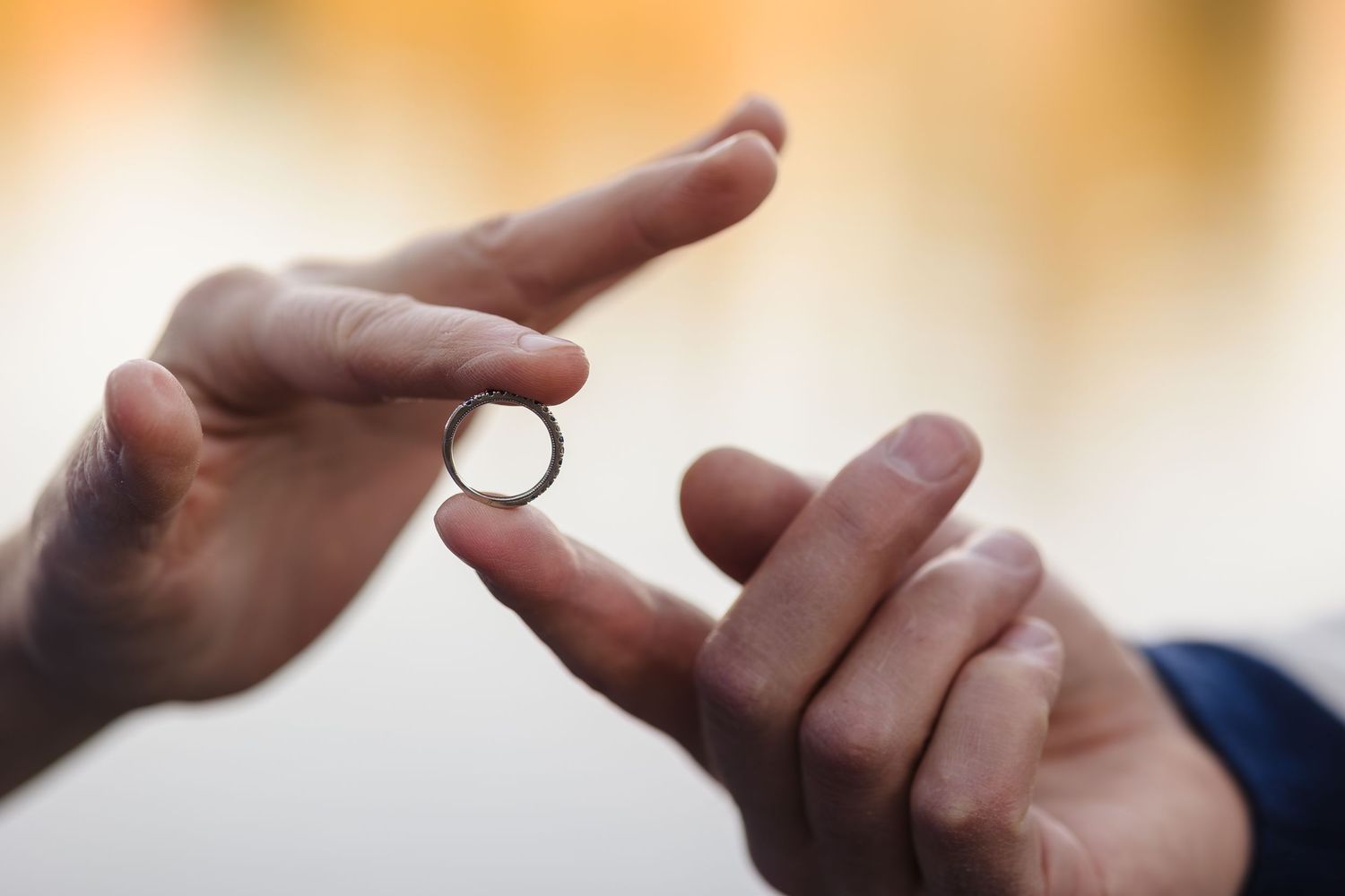 Hands exchanging ring — Close‑up of two hands exchanging a silver wedding ring with a warm, glowing bokeh background.