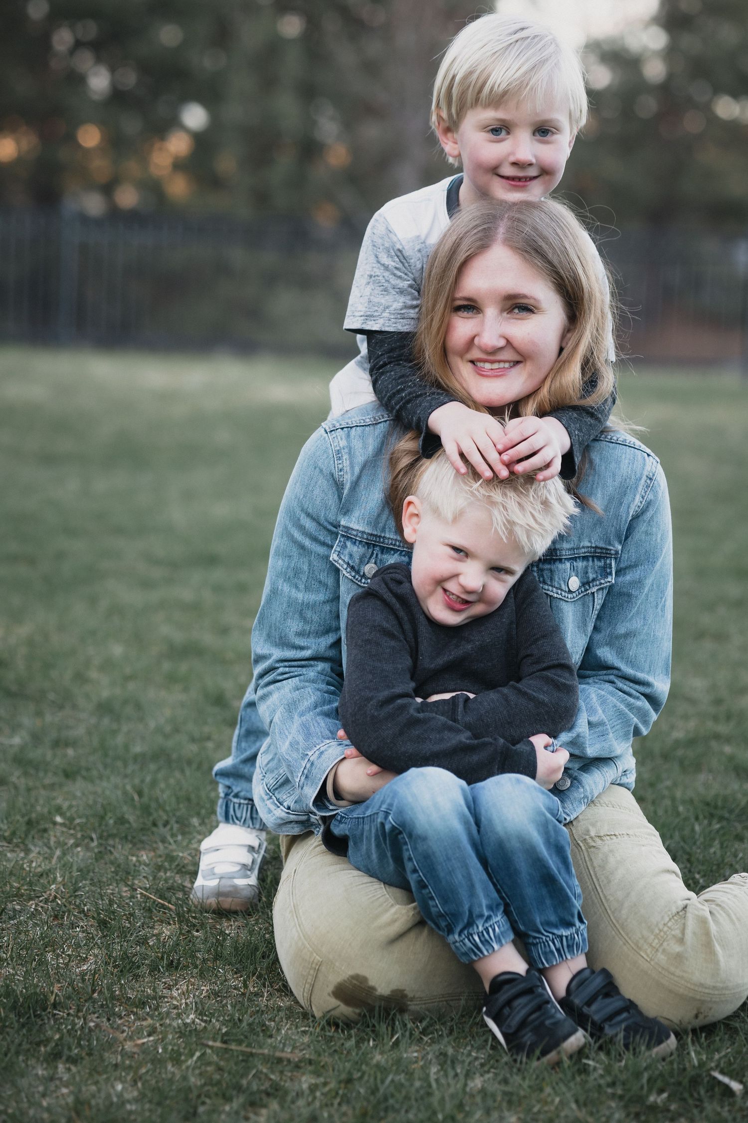 Mom with boys hugging — Smiling mother in a denim jacket sitting on the grass as her two blonde boys hug her warmly.