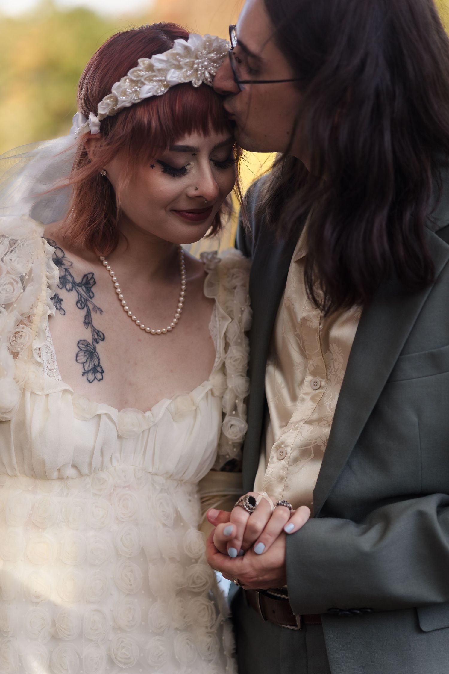 Gothic bride with red hair and floral headpiece receives kiss from groom in gray suit at outdoor wedding.