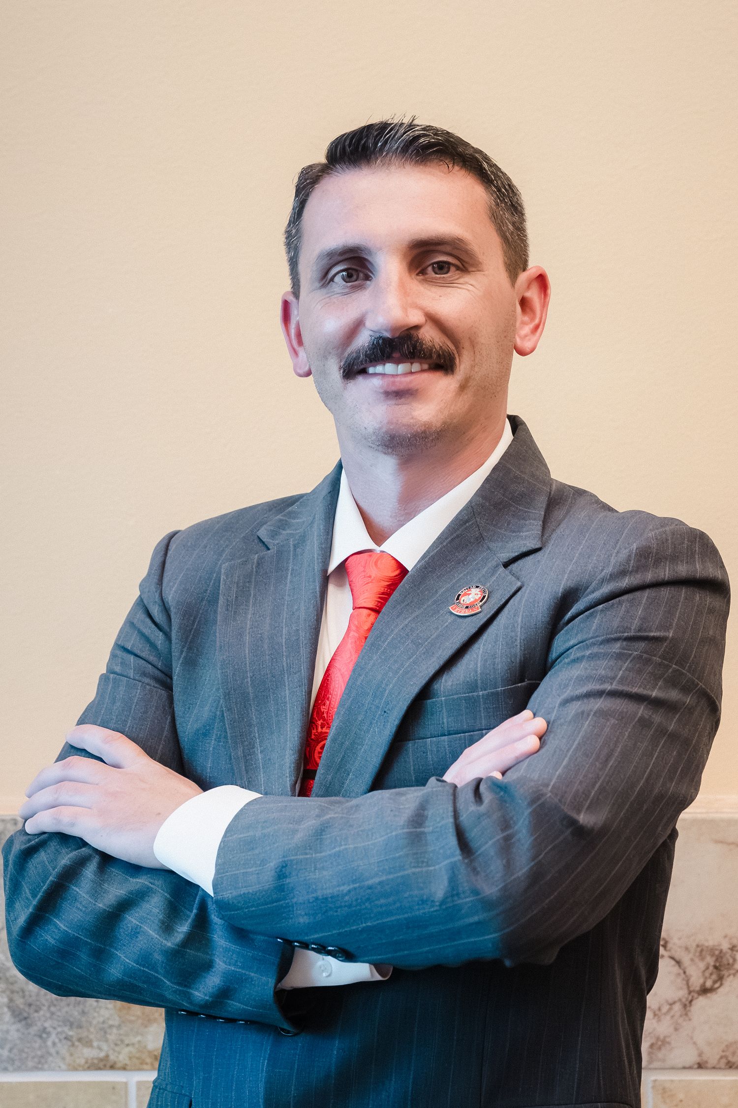 Professional man in gray pinstripe suit and red tie smiling with arms crossed against neutral background.