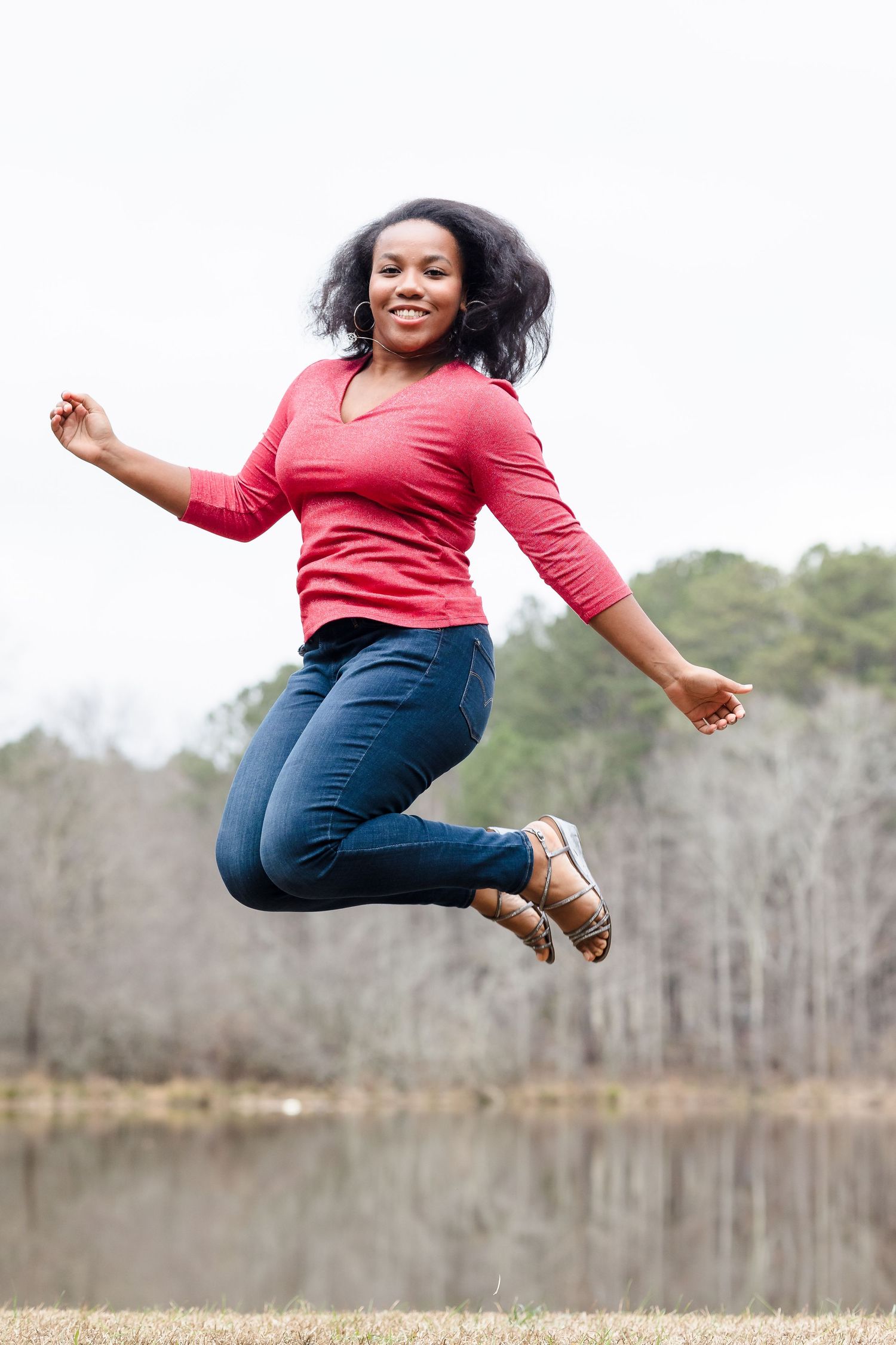 Joyful woman jumping by lake — Joyful Black woman jumping mid‑air in a red top and jeans near a lake with bare winter trees.