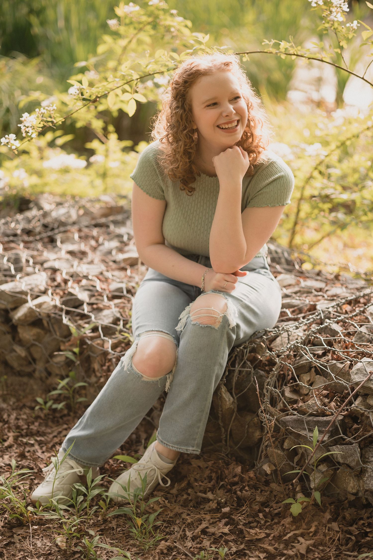 Curly red-haired girl sitting on rocks in a sunlit garden, wearing mint top and ripped jeans, laughing.