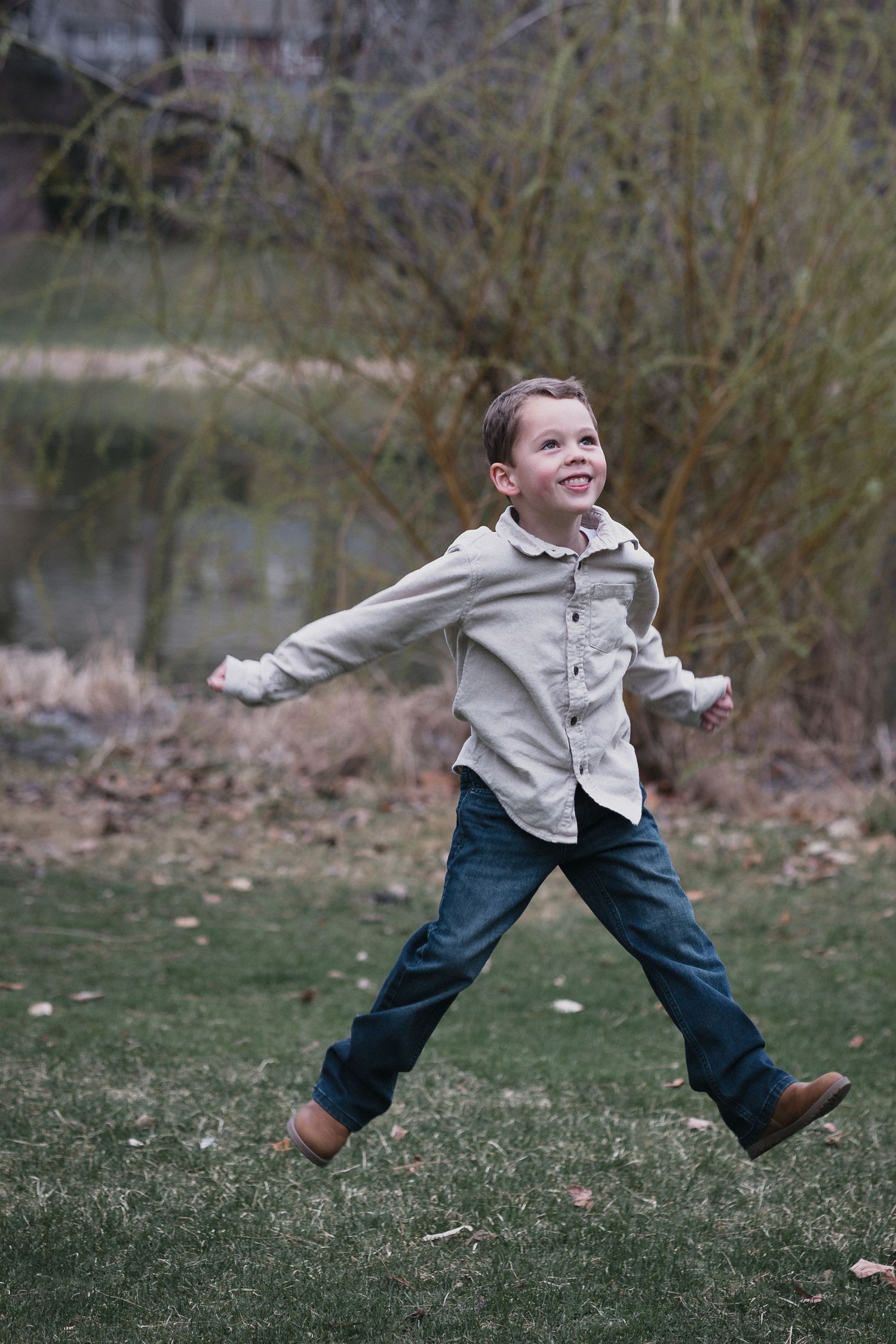 Boy jumping by pond — Happy young boy in a beige shirt jumping playfully in a park near a pond.