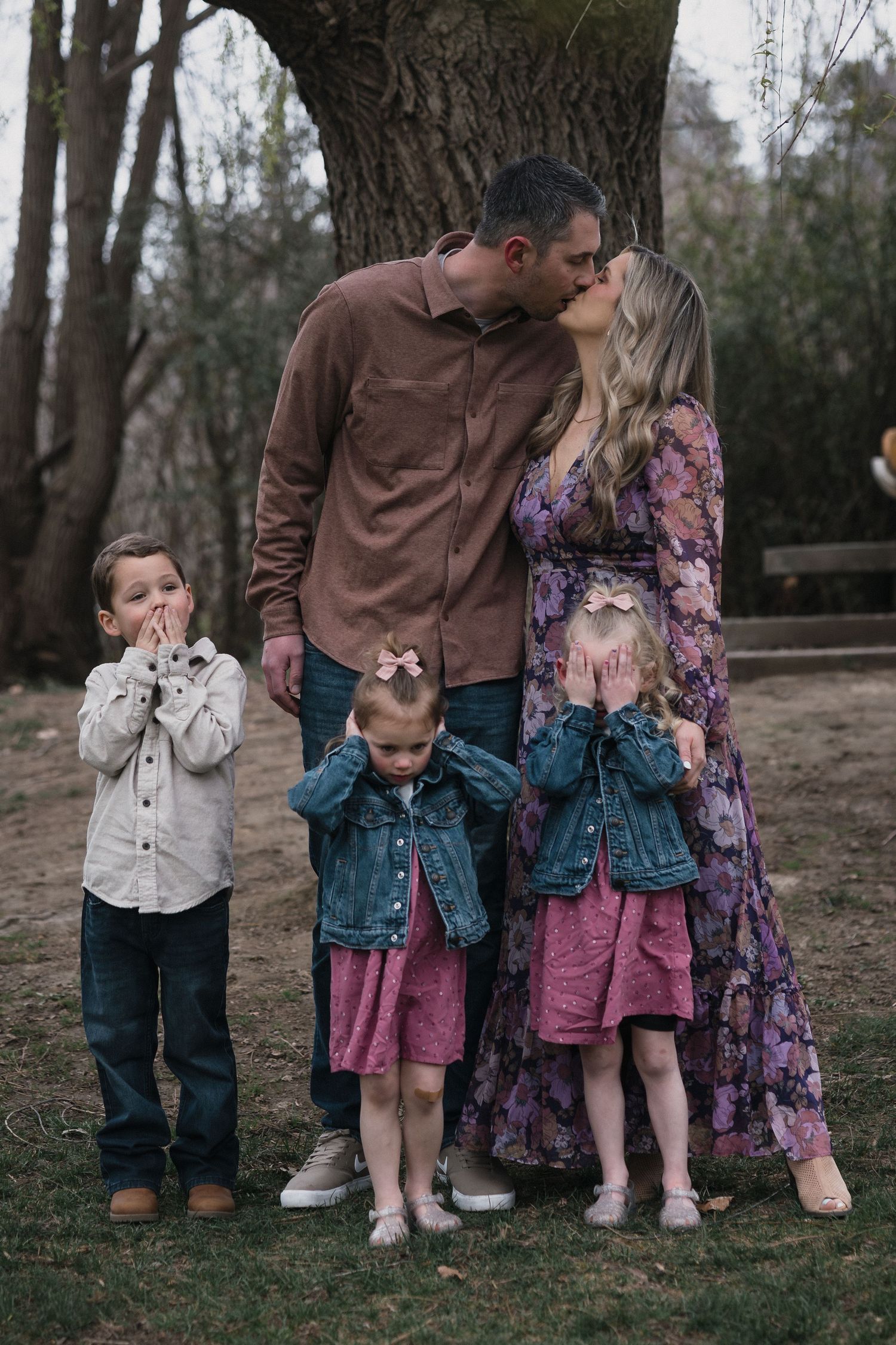 Family of five playful pose — Family of five outdoors with parents kissing while three children cover their eyes in a playful moment.