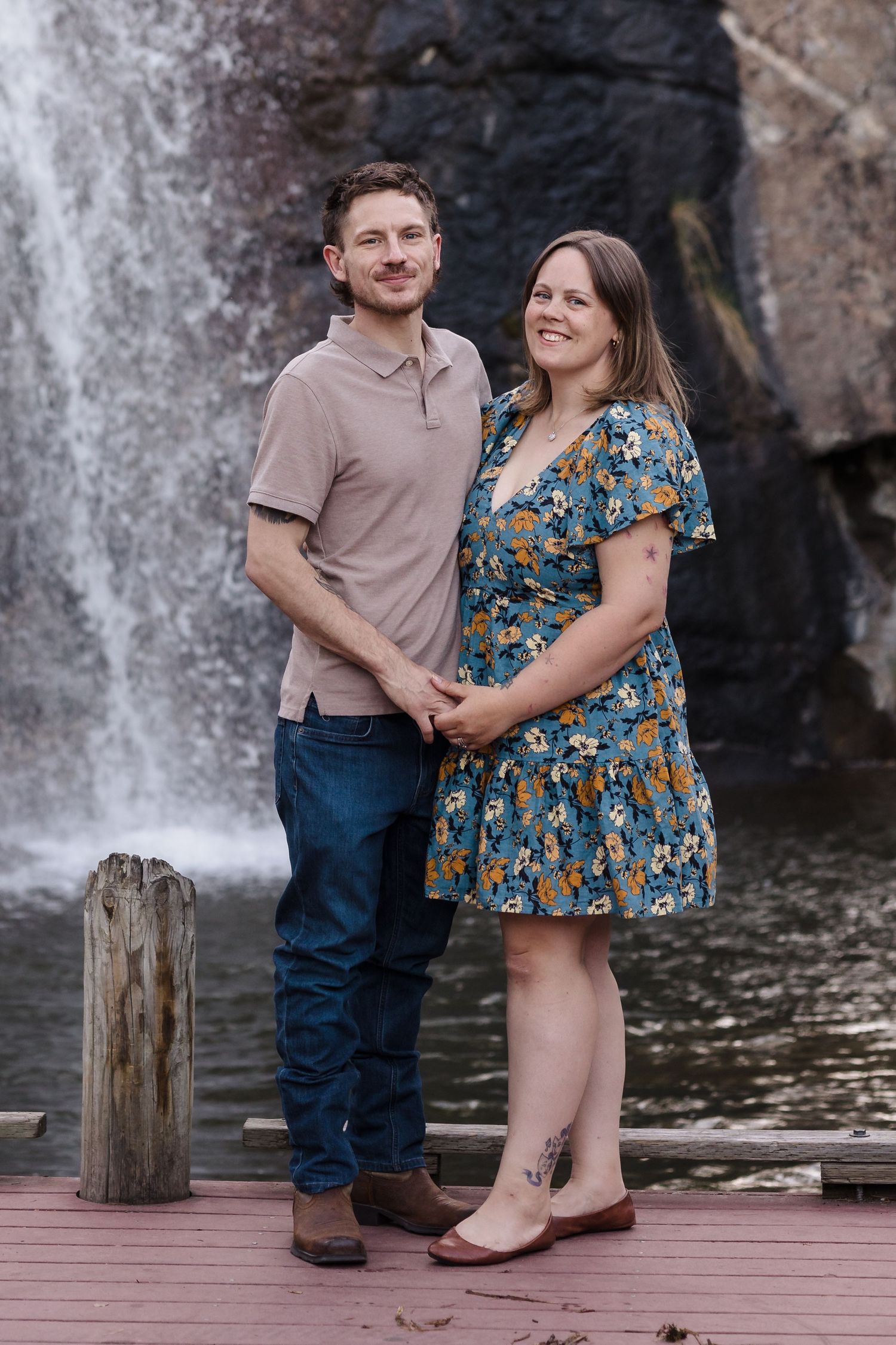 Couple by waterfall deck — Couple posing on a wooden deck in front of a waterfall, he in a beige polo, she in a blue floral dress.