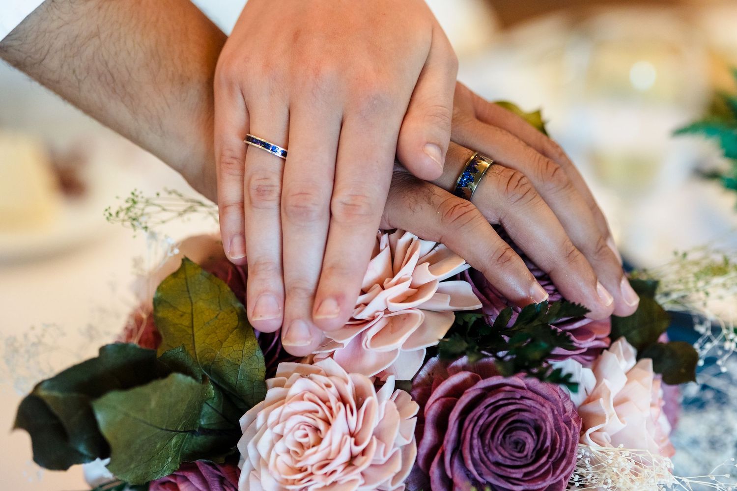Newlywed couple placing hands on pink and purple rose wedding bouquet showing matching gold bands.