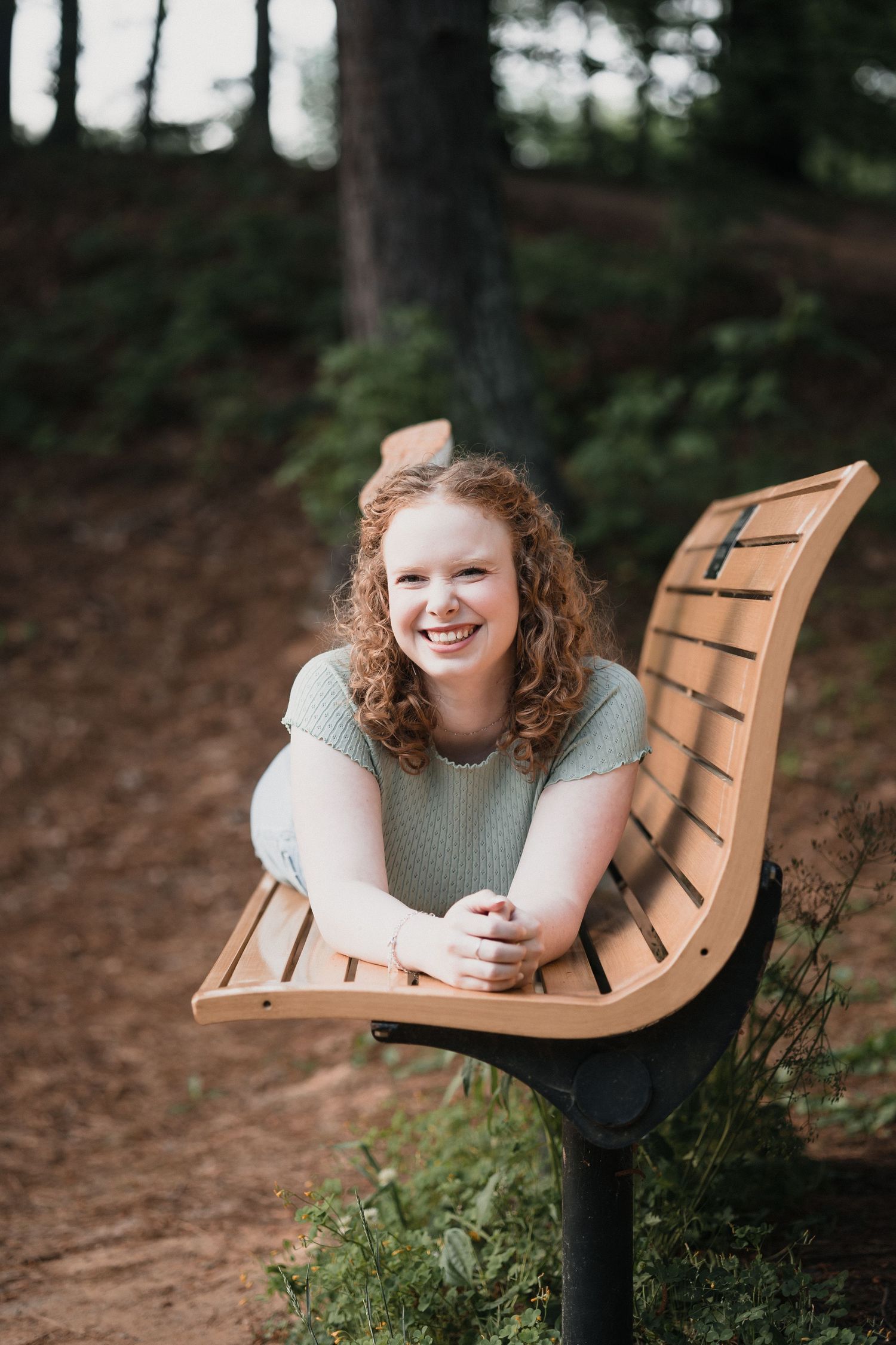Red‑haired woman on bench — Warm outdoor portrait of smiling young woman with curly red hair leaning on a wooden park bench
