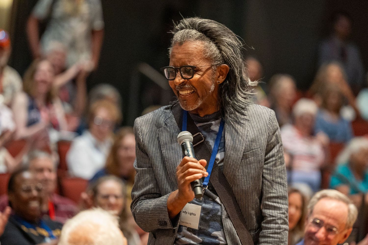 A smiling woman with dreadlocks in a gray blazer holds a microphone at an indoor event.
