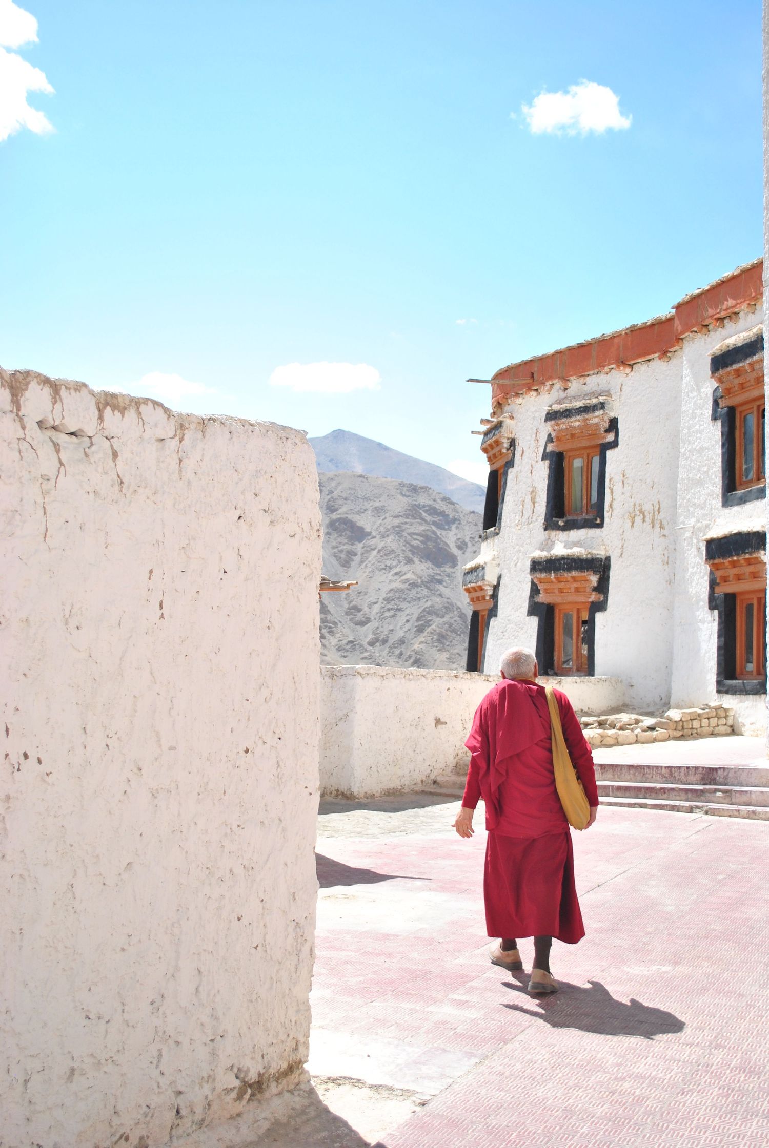 A Buddhist monk in red robes walks through a Tibetan monastery courtyard with white walls and mountains in the background.