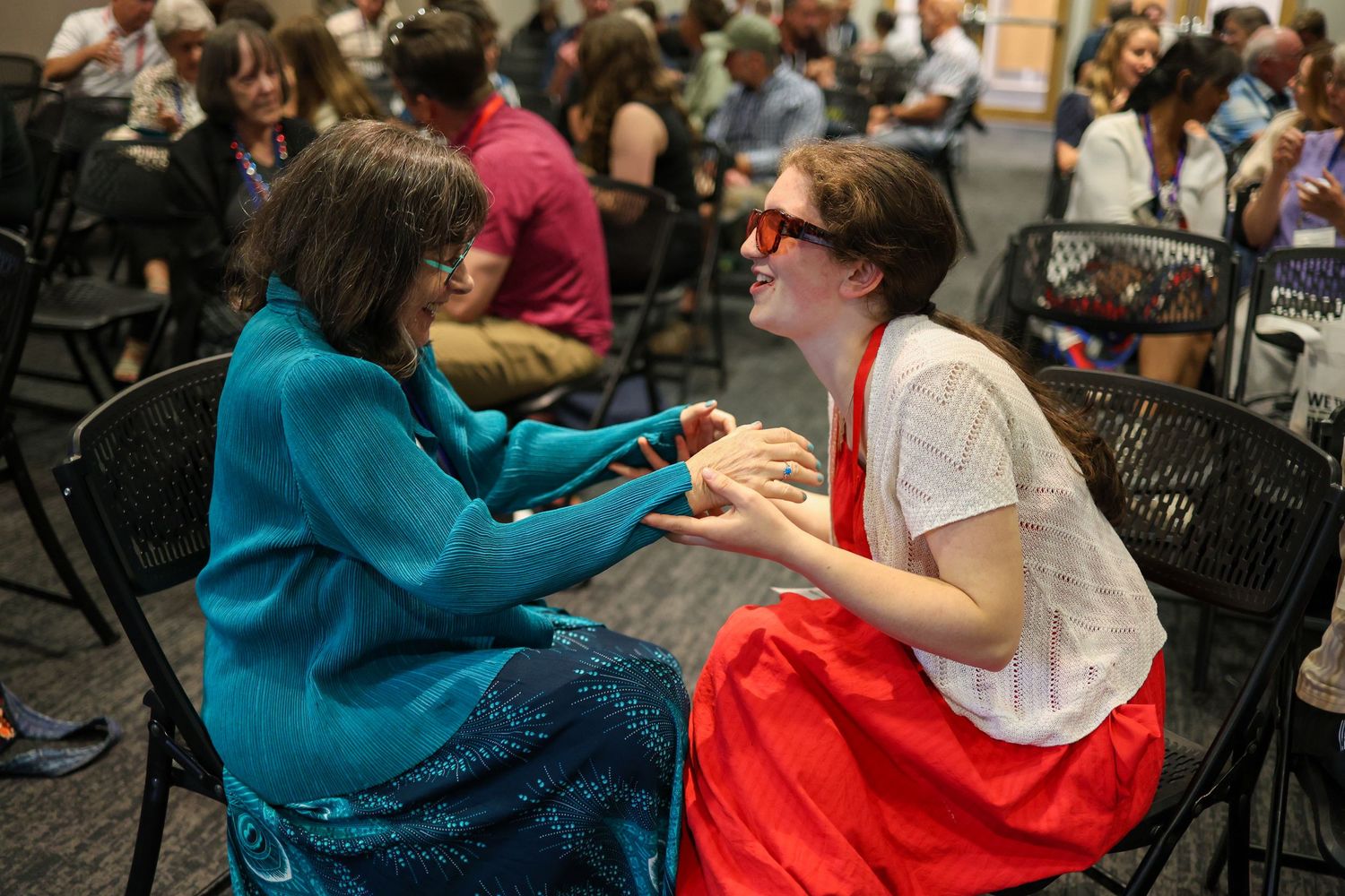 Two women share a joyful conversation while seated on black chairs at a busy indoor event.
