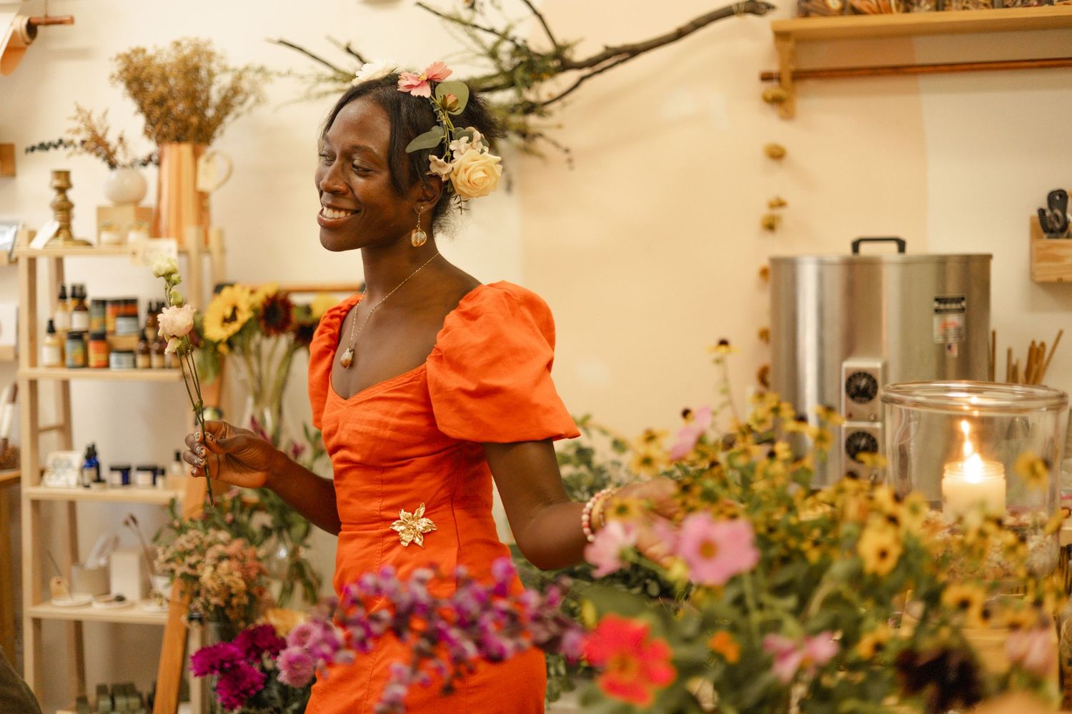 Woman in orange dress arranging colorful flowers in a floral shop, smiling joyfully with blooms in her hair.