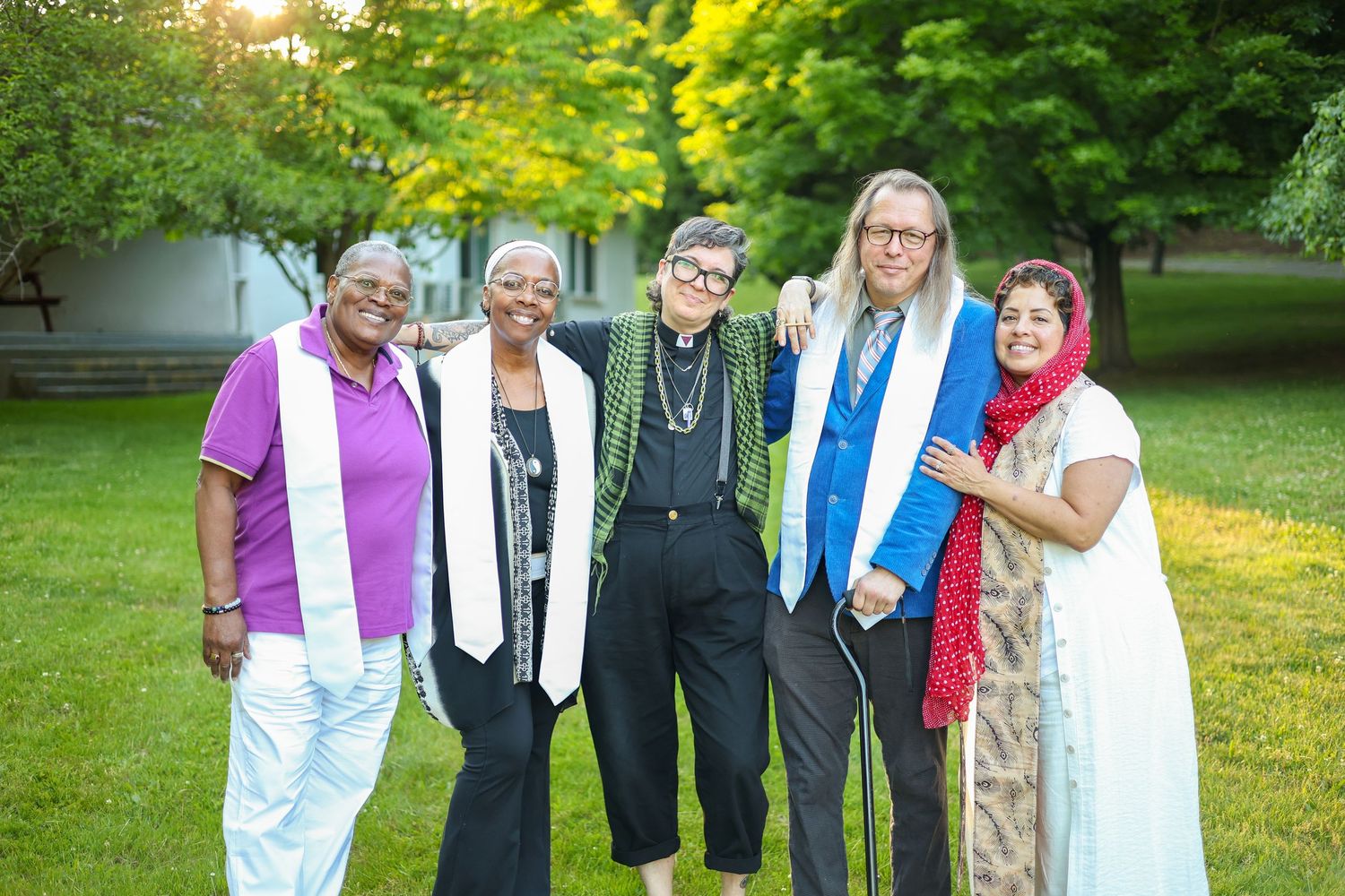 Five diverse women smiling outdoors at a graduation ceremony, wearing stoles and colorful attire on a green lawn.