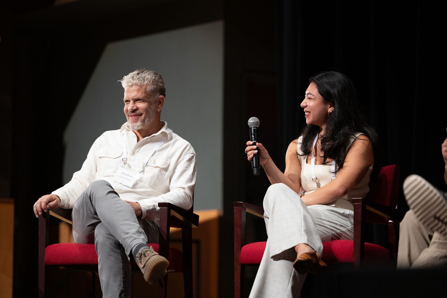 Two people in white outfits sit on stage during a panel discussion, woman holds microphone while man smiles.
