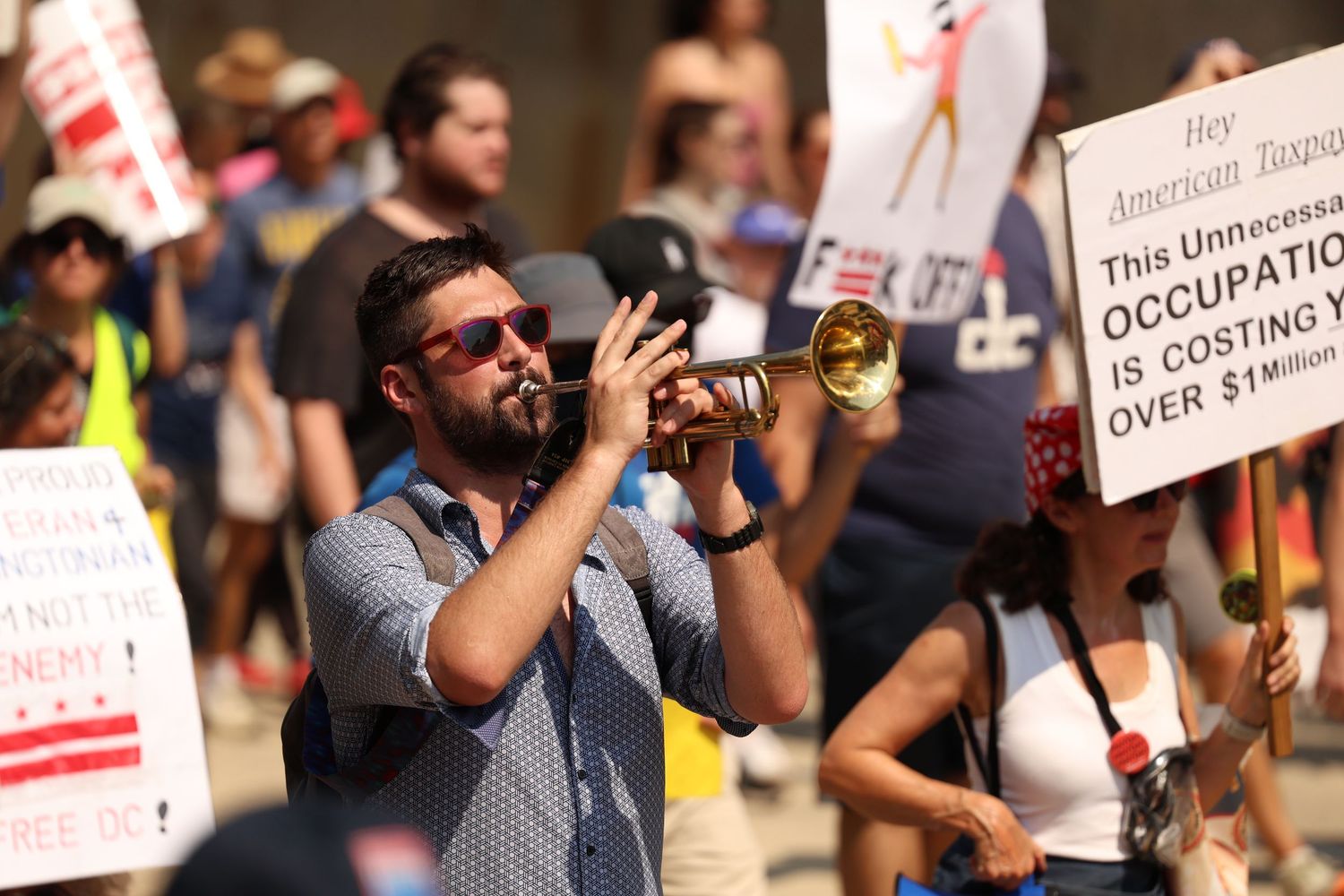 Bearded man plays trumpet at an outdoor protest with signs and crowd behind him.