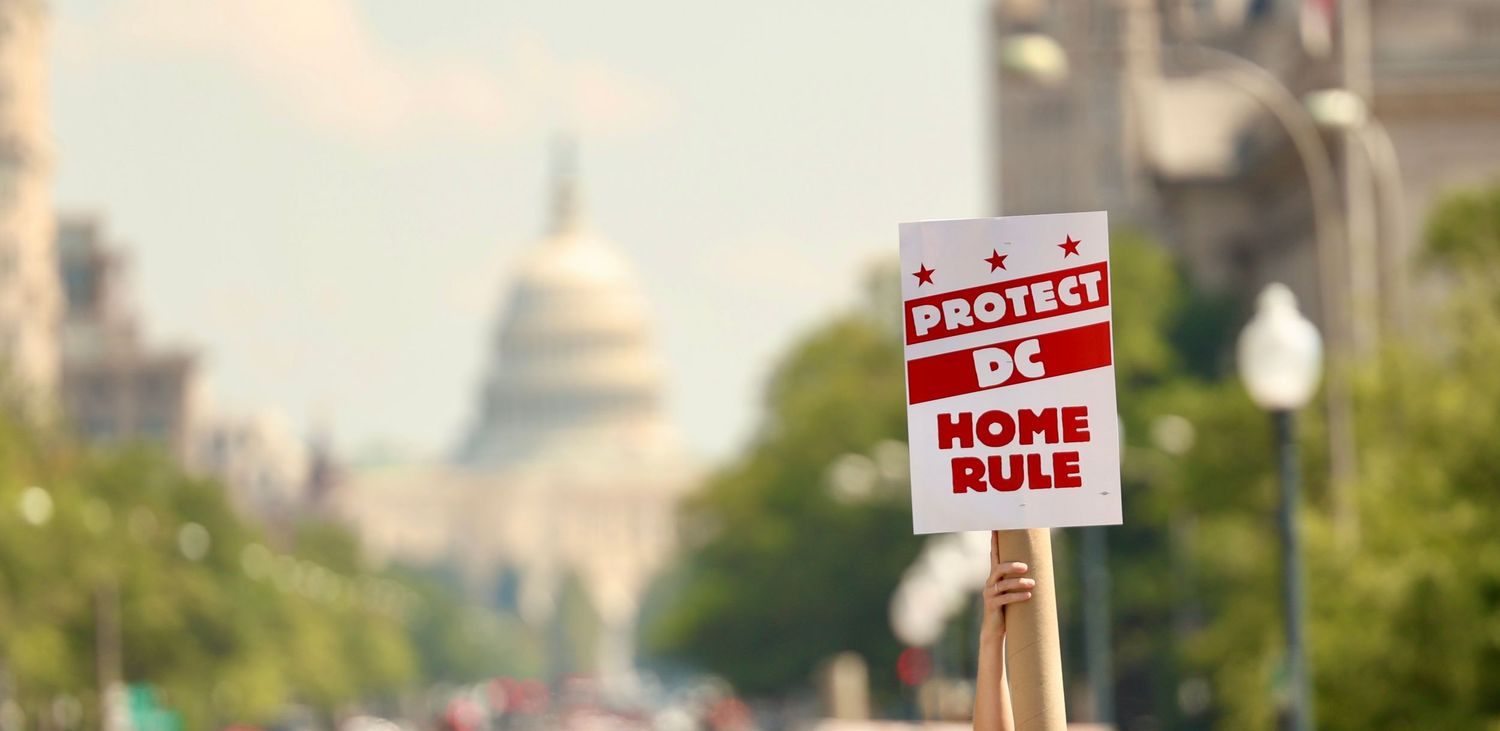A hand holds a Protect DC Home Rule sign with the US Capitol building visible in the background.