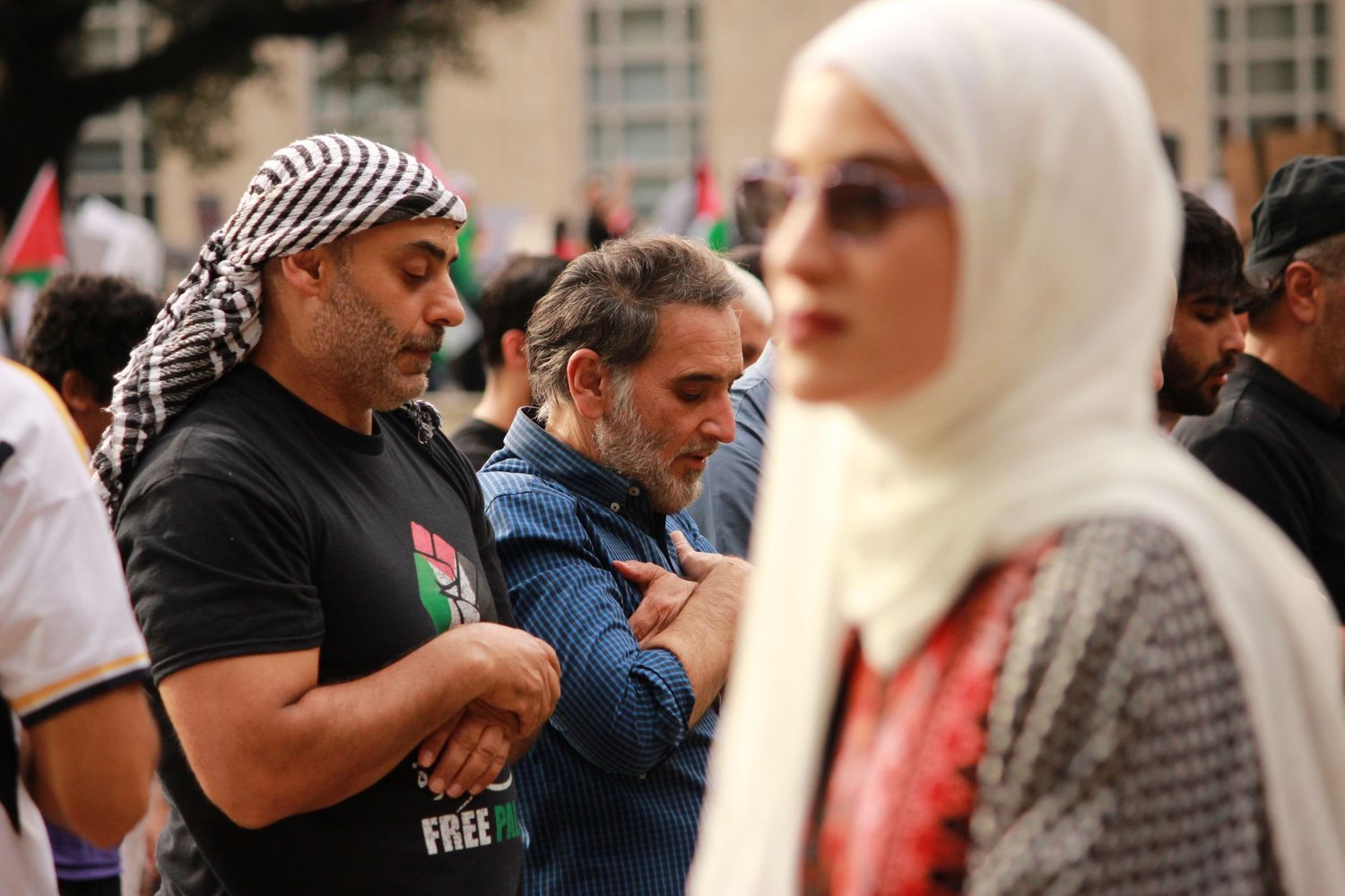 Two men pray at a rally as a woman in a hijab stands nearby.