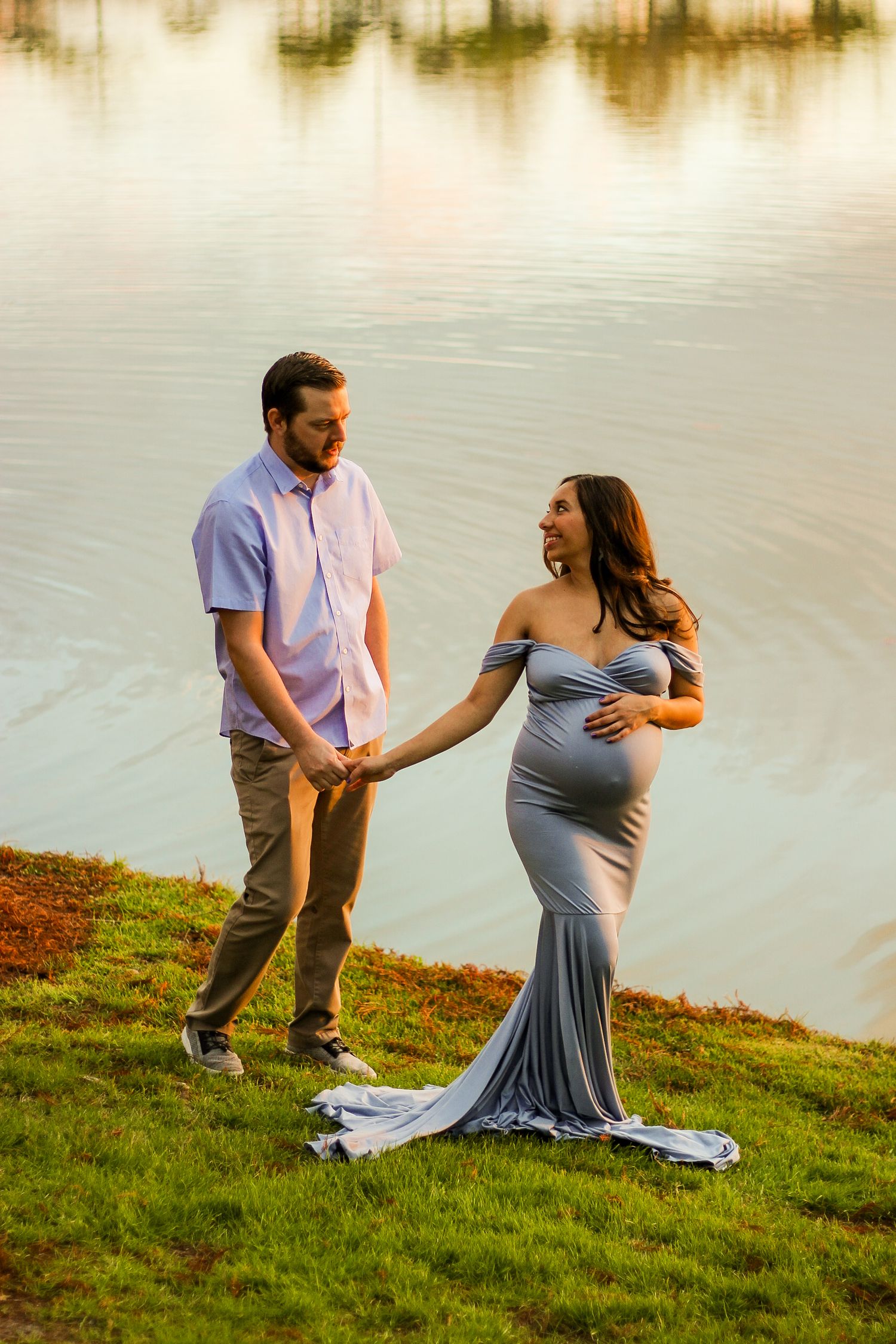 Pregnant woman in gray gown holds hands with man by a lake during a golden hour maternity photoshoot.
