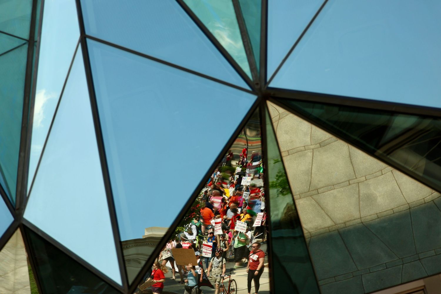 Colorful crowd reflected in geometric triangular glass panels of a modern building facade under blue sky.