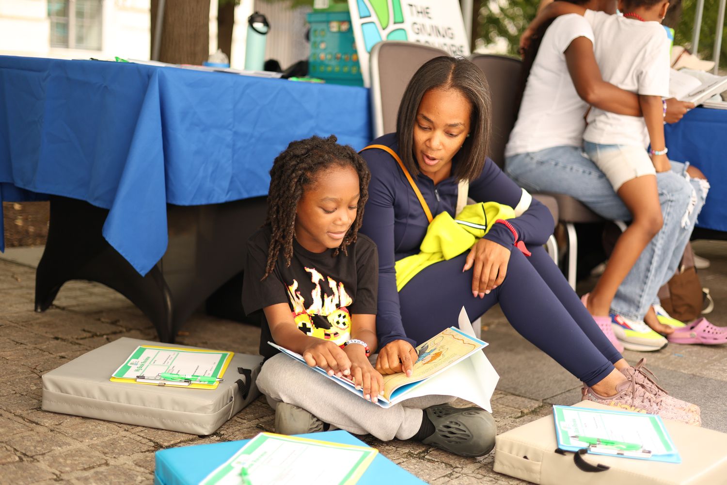 A woman and young girl sit together reading a book at an outdoor community literacy event.