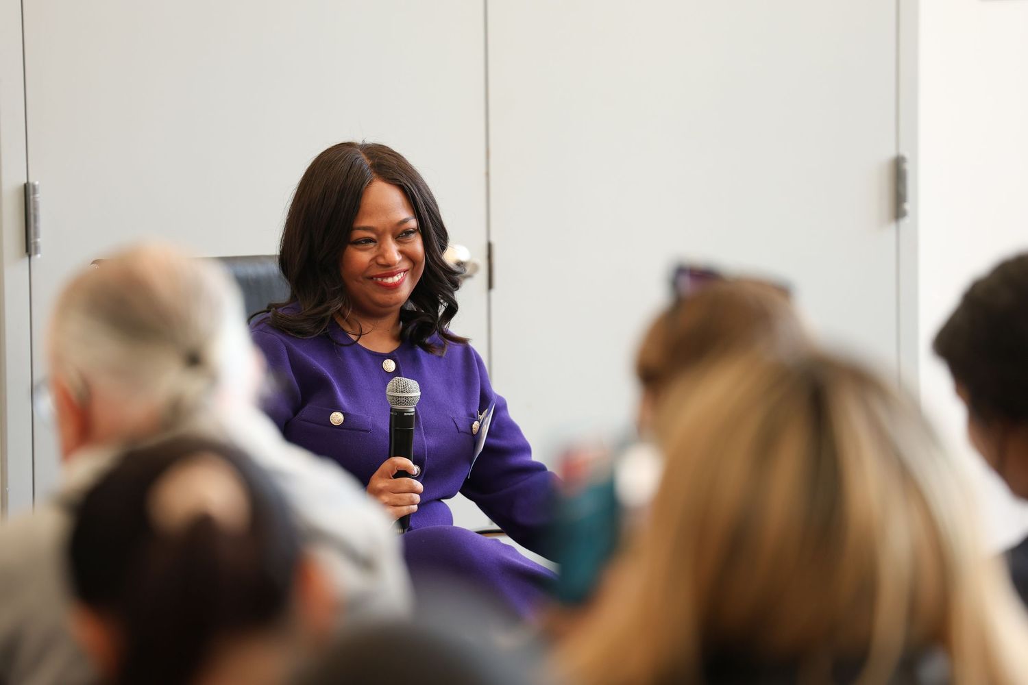 Woman in purple blazer speaking into microphone at panel discussion, smiling at audience.